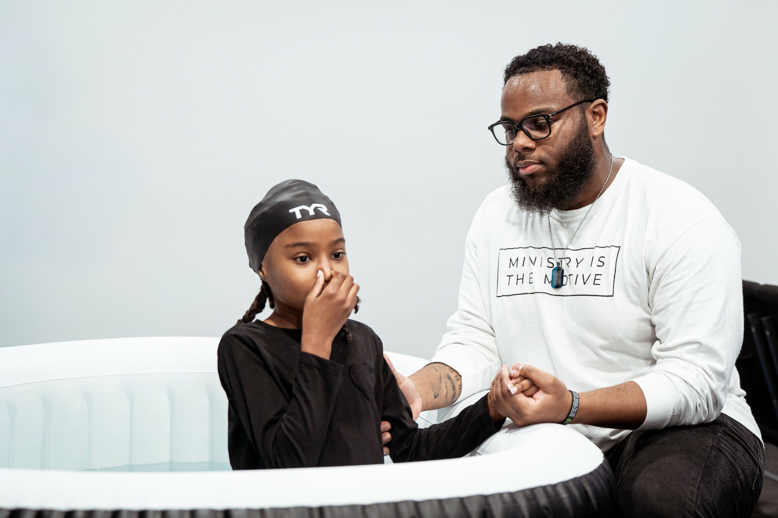 Adult man and young girl sitting in a white circular seating area, holding hands, with the girl covering her mouth.