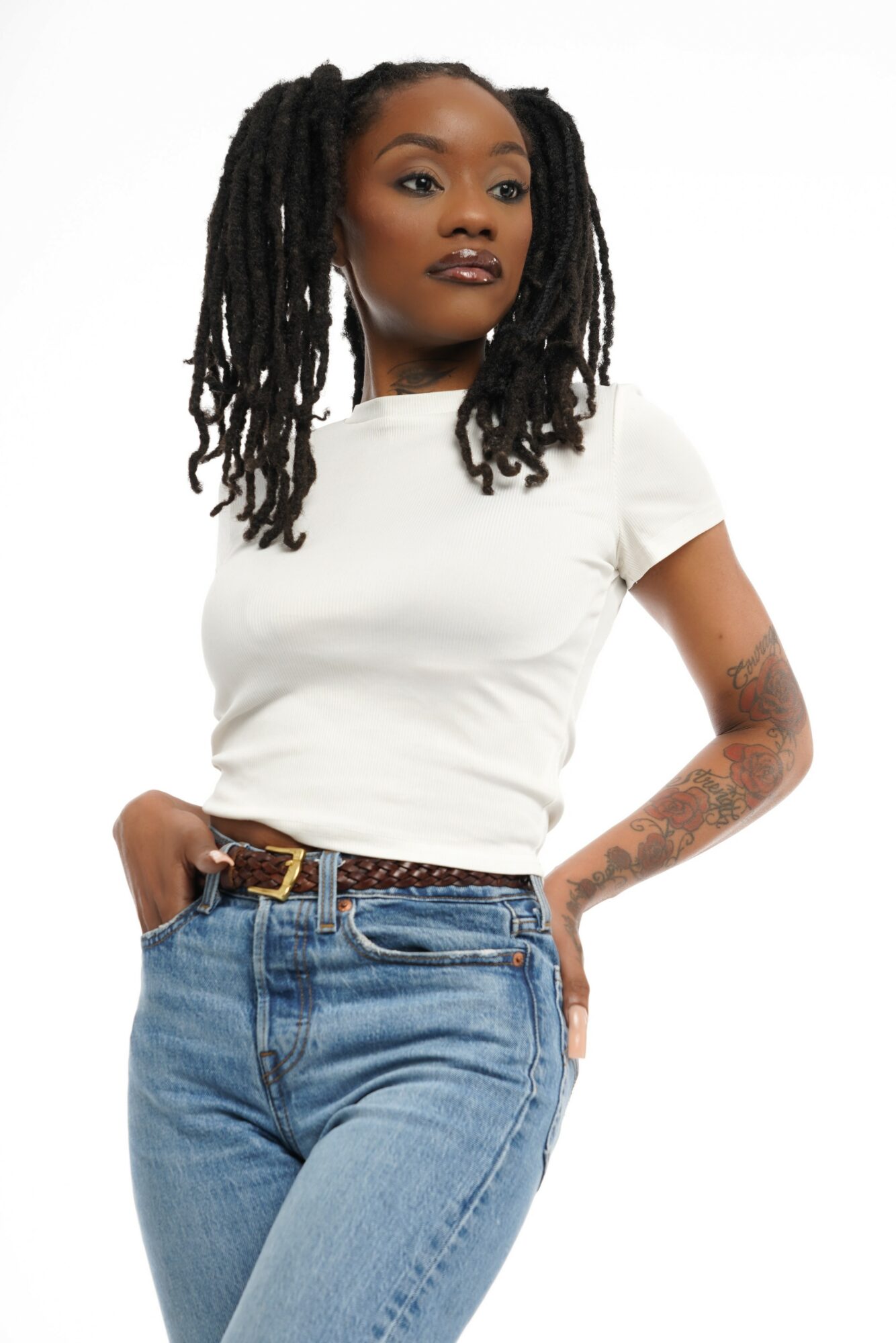 Woman with dreadlocks wearing a white t-shirt and jeans, standing with hands in pockets.
