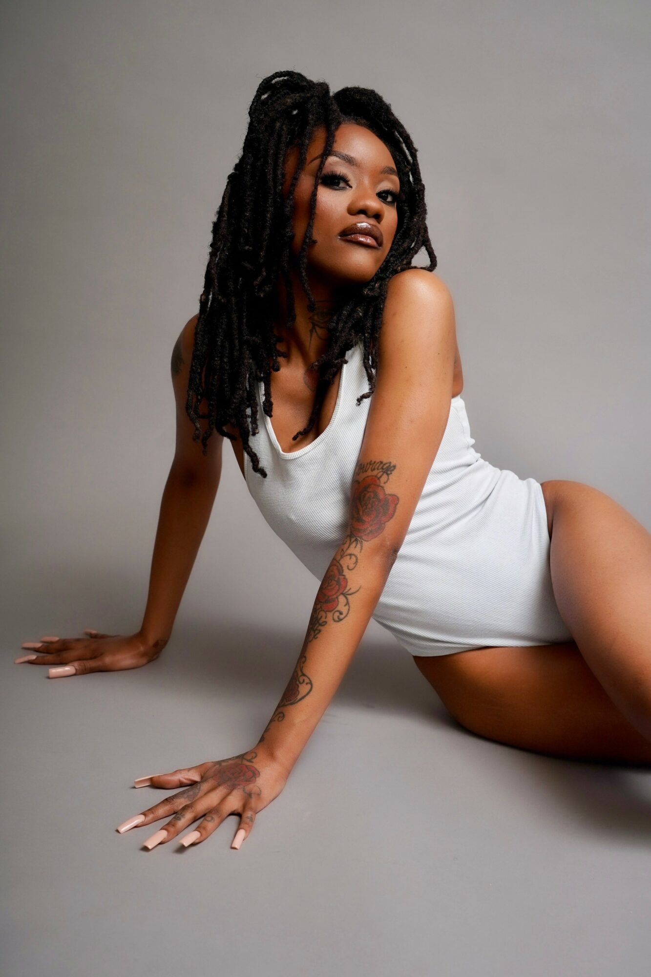 Woman with dreadlocks in a white dress, sitting on the floor, looking at the camera.