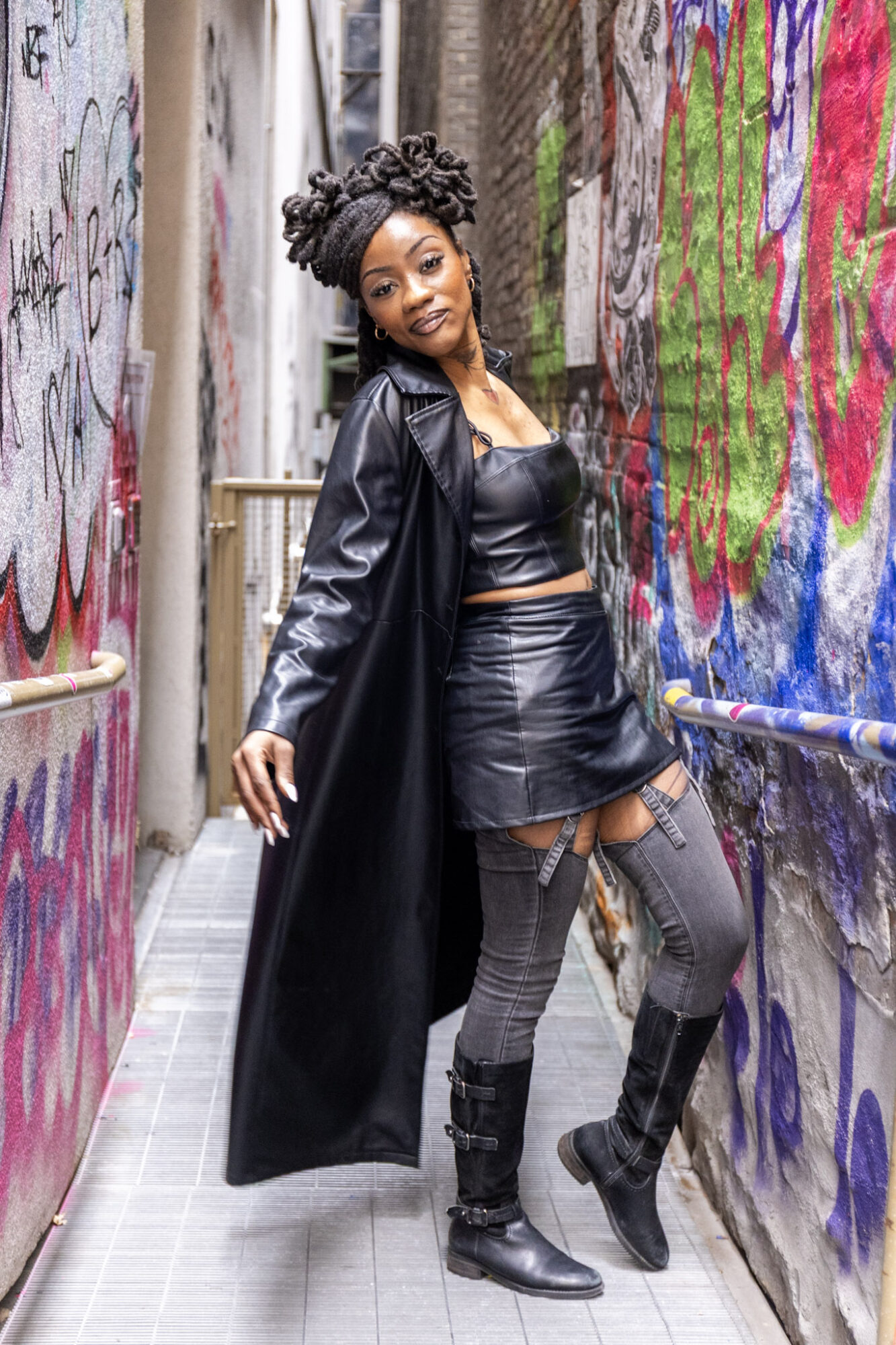 Young woman with curly hair in leather outfit and long coat standing in graffiti-covered alleyway.