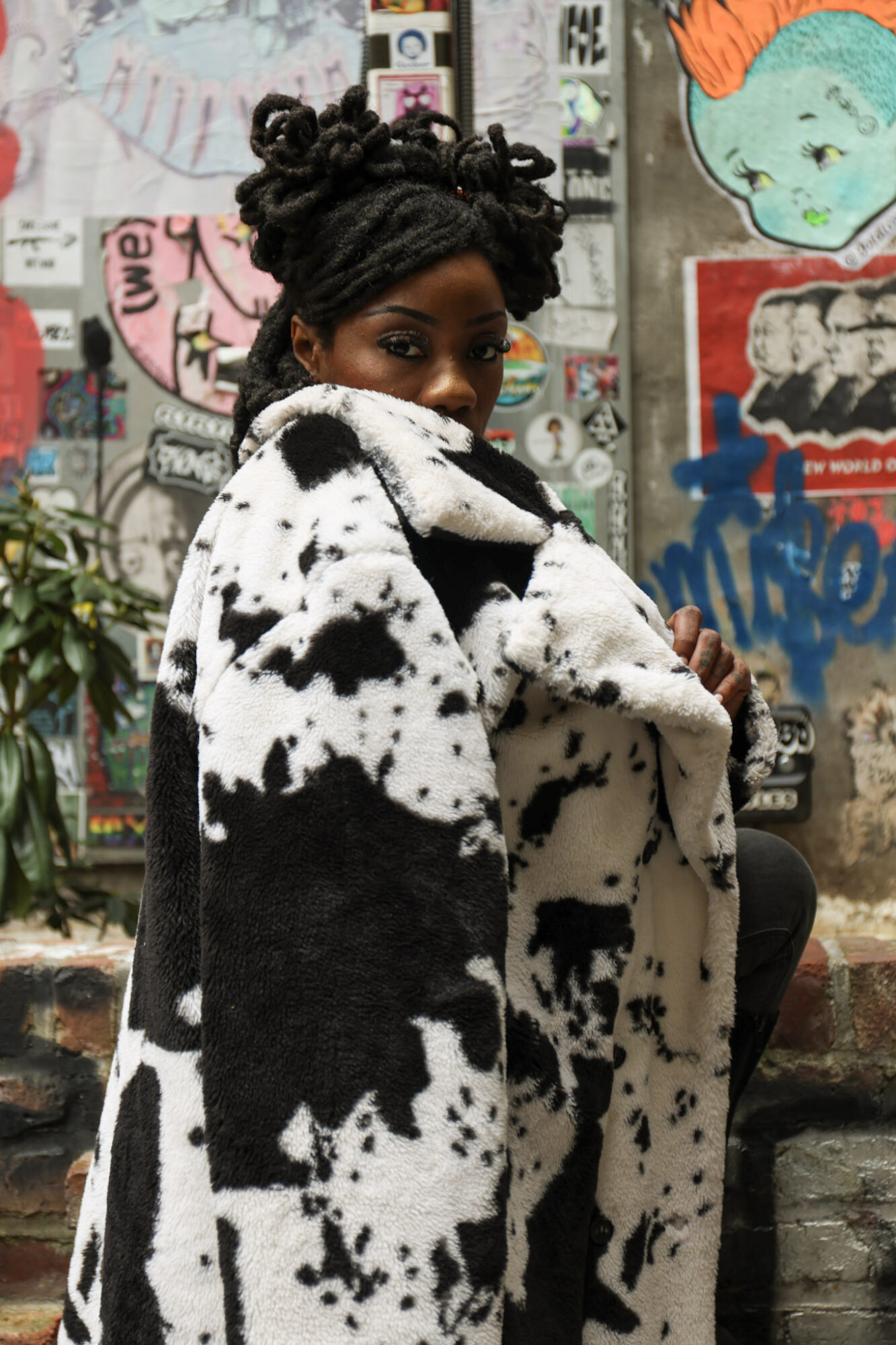 Woman with styled hair wearing a black and white patterned coat, standing in front of graffiti-covered wall.