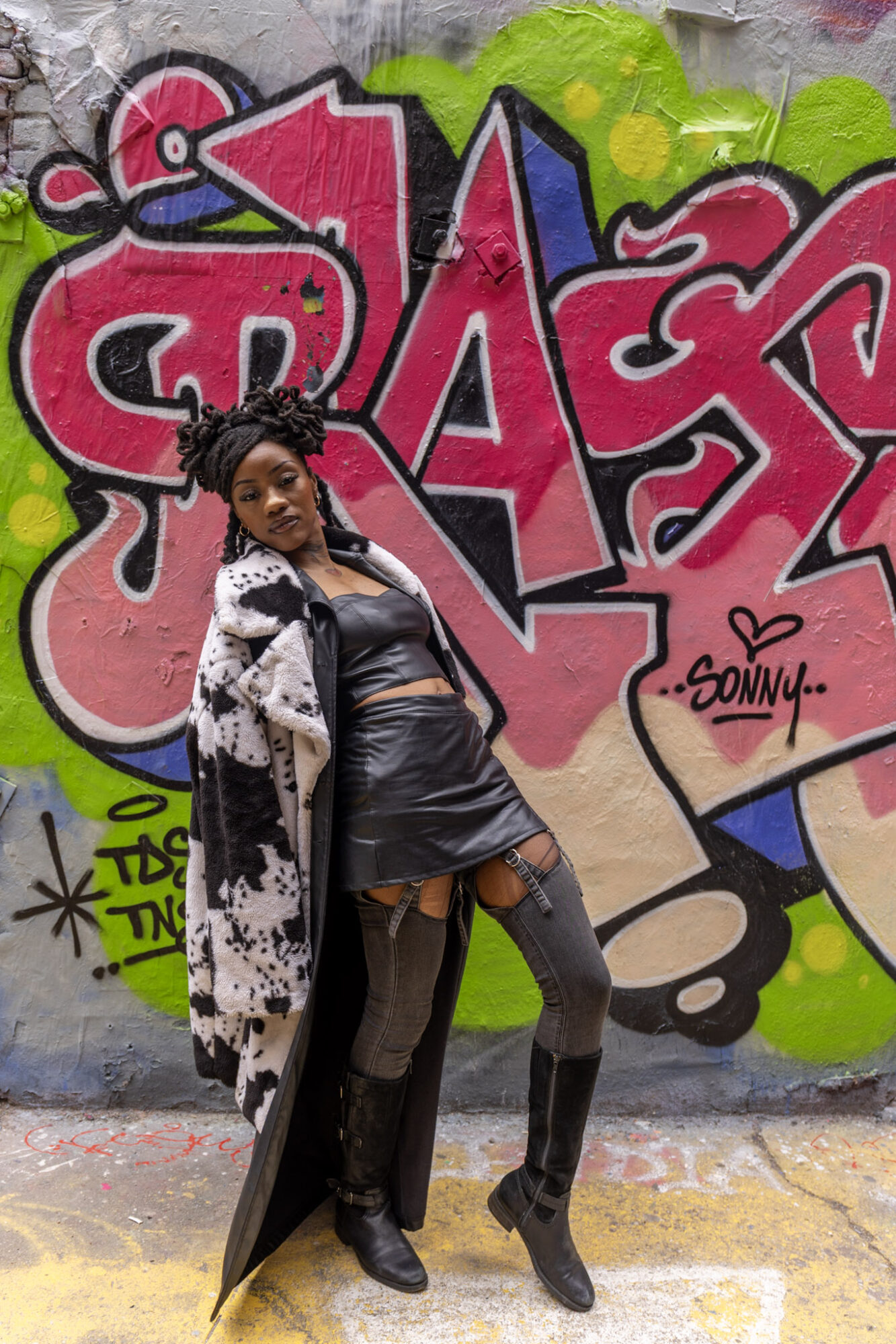 Woman standing in front of colorful graffiti wall, wearing a black dress and long coat, with one hand on her hip.