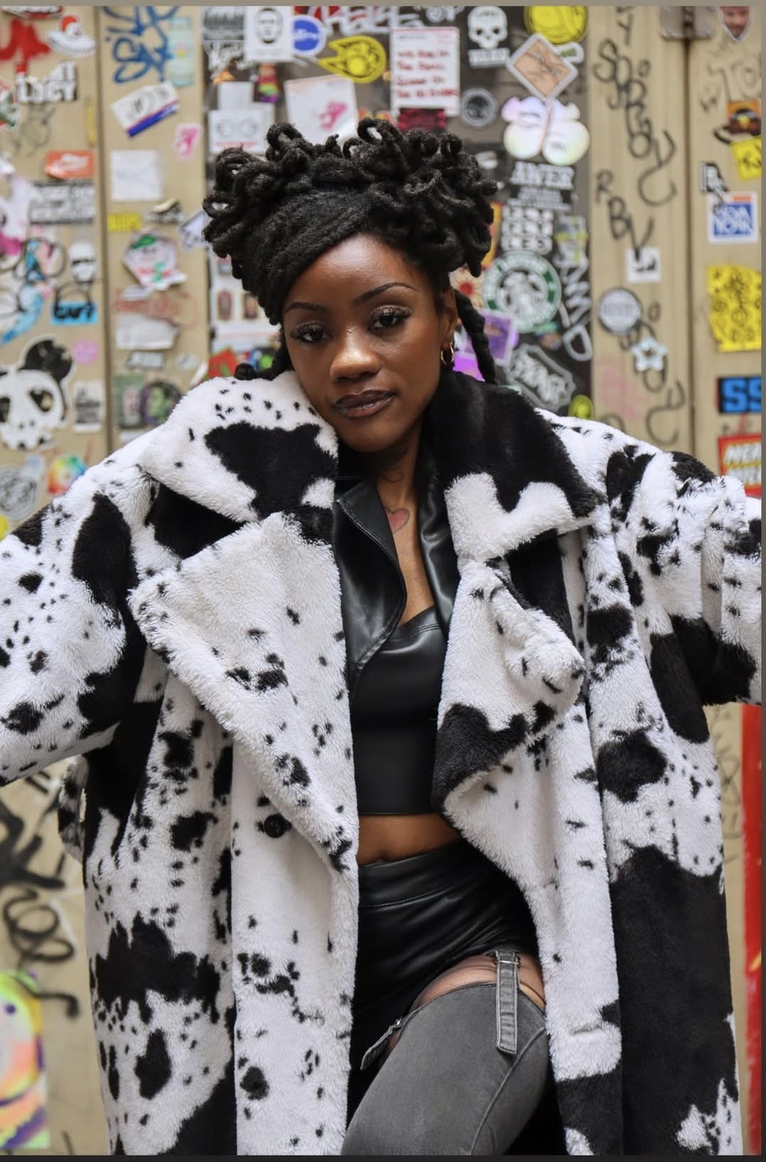 Woman with braided hair wearing a black top and a black-and-white spotted coat, standing in front of a colorful wall.