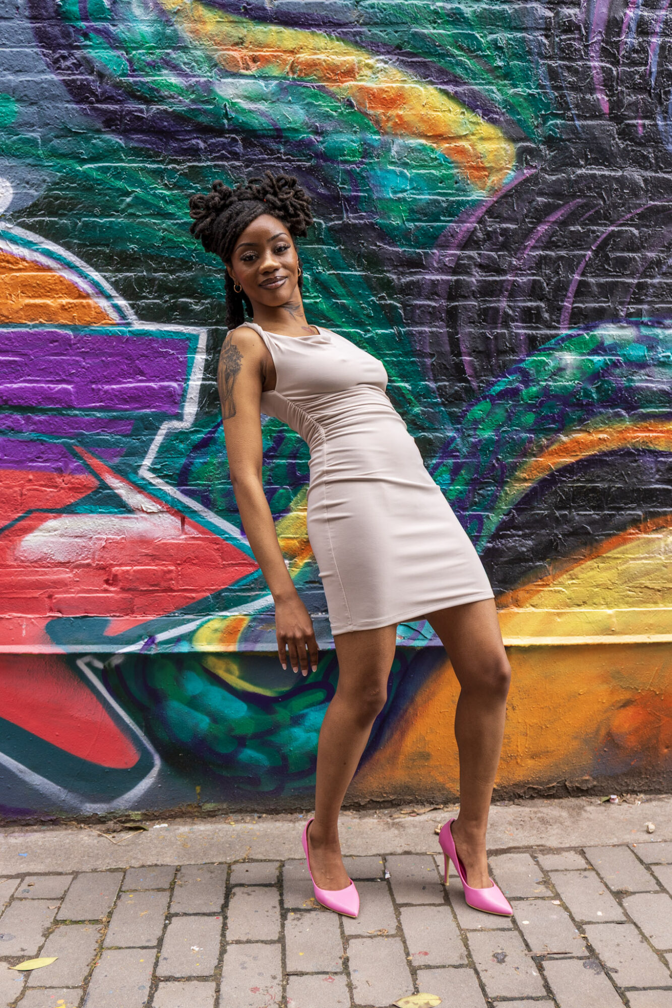 Woman in a beige dress and pink heels standing in front of colorful graffiti wall.
