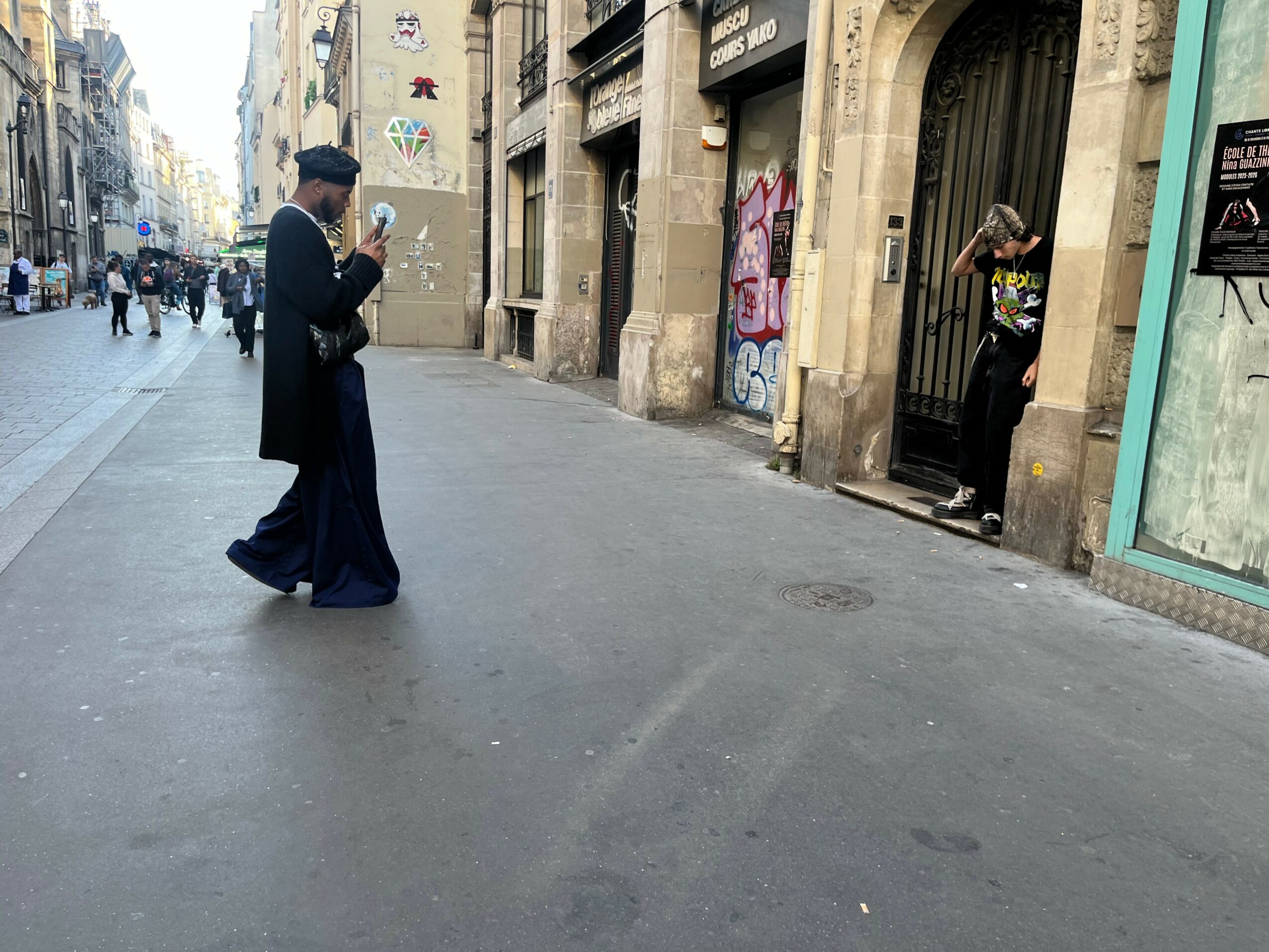 Two men on a city street, one looking at a phone and the other standing near a doorway, with buildings and graffiti.