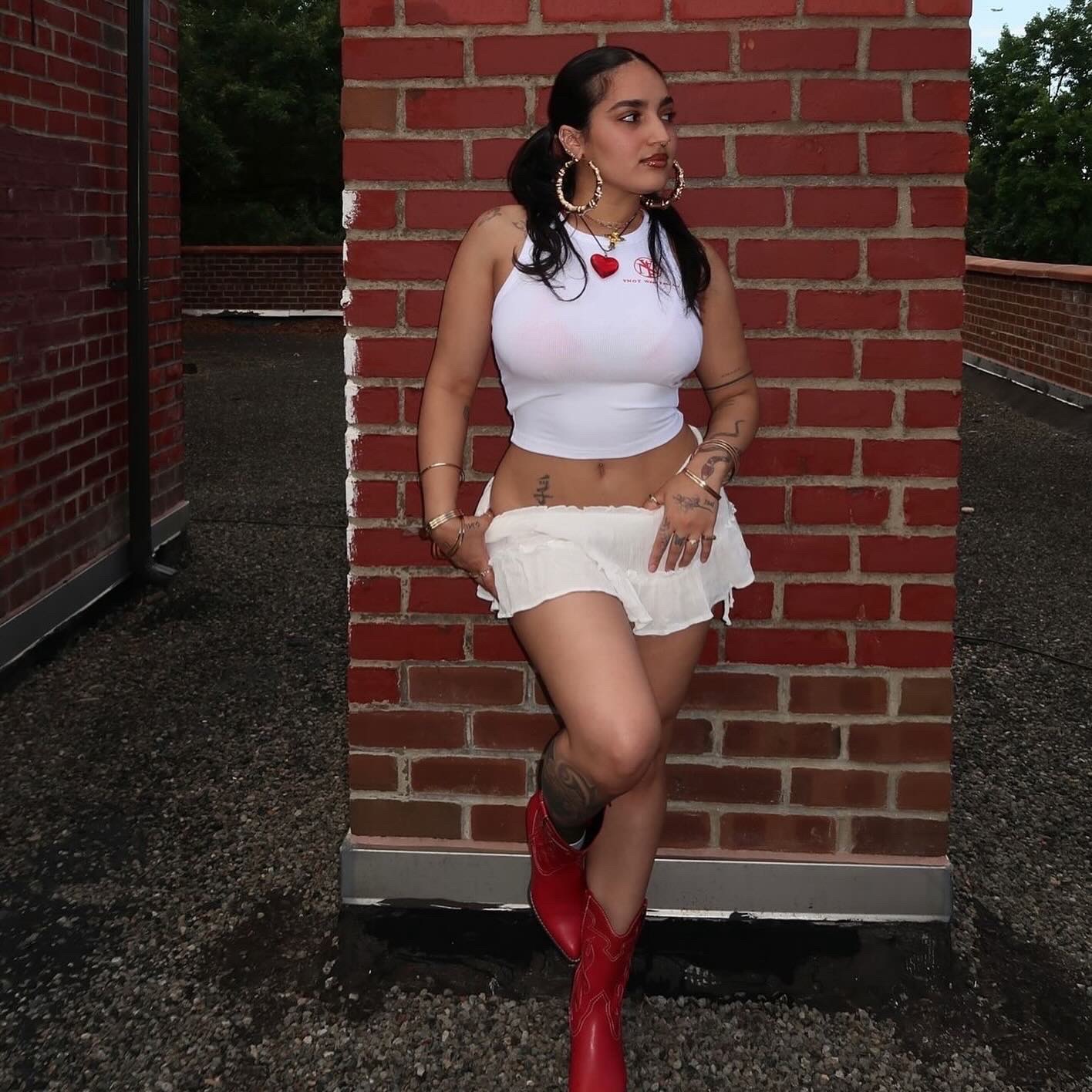 Woman in white crop top and shorts with red boots standing against a brick wall outdoors.