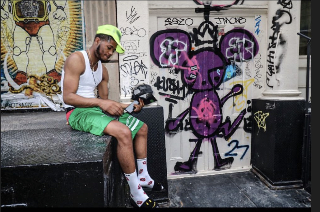 Man sitting on a black block, wearing a white tank top, green shorts, and a green cap, with graffiti art behind him.