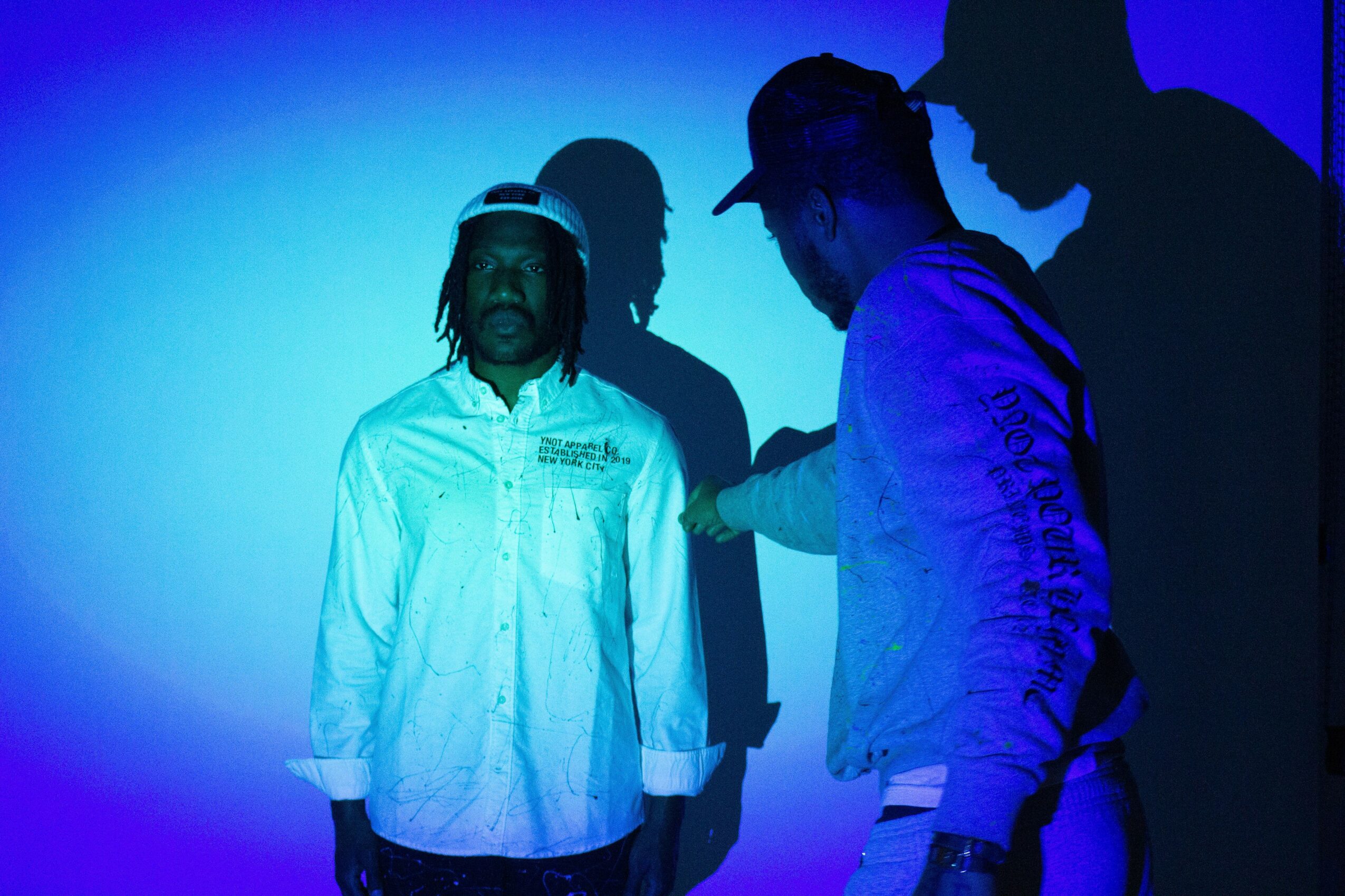 Two people in casual clothing and hats stand against a blue-lit background, casting shadows on the wall behind them.