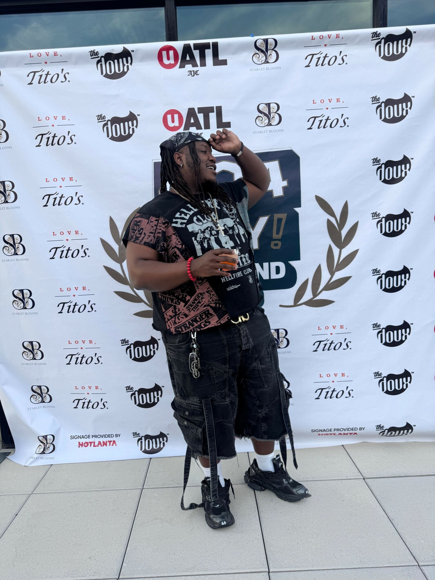 Young man with dreadlocks and tattoos standing in front of a promotional backdrop, holding a drink, wearing casual clothes.
