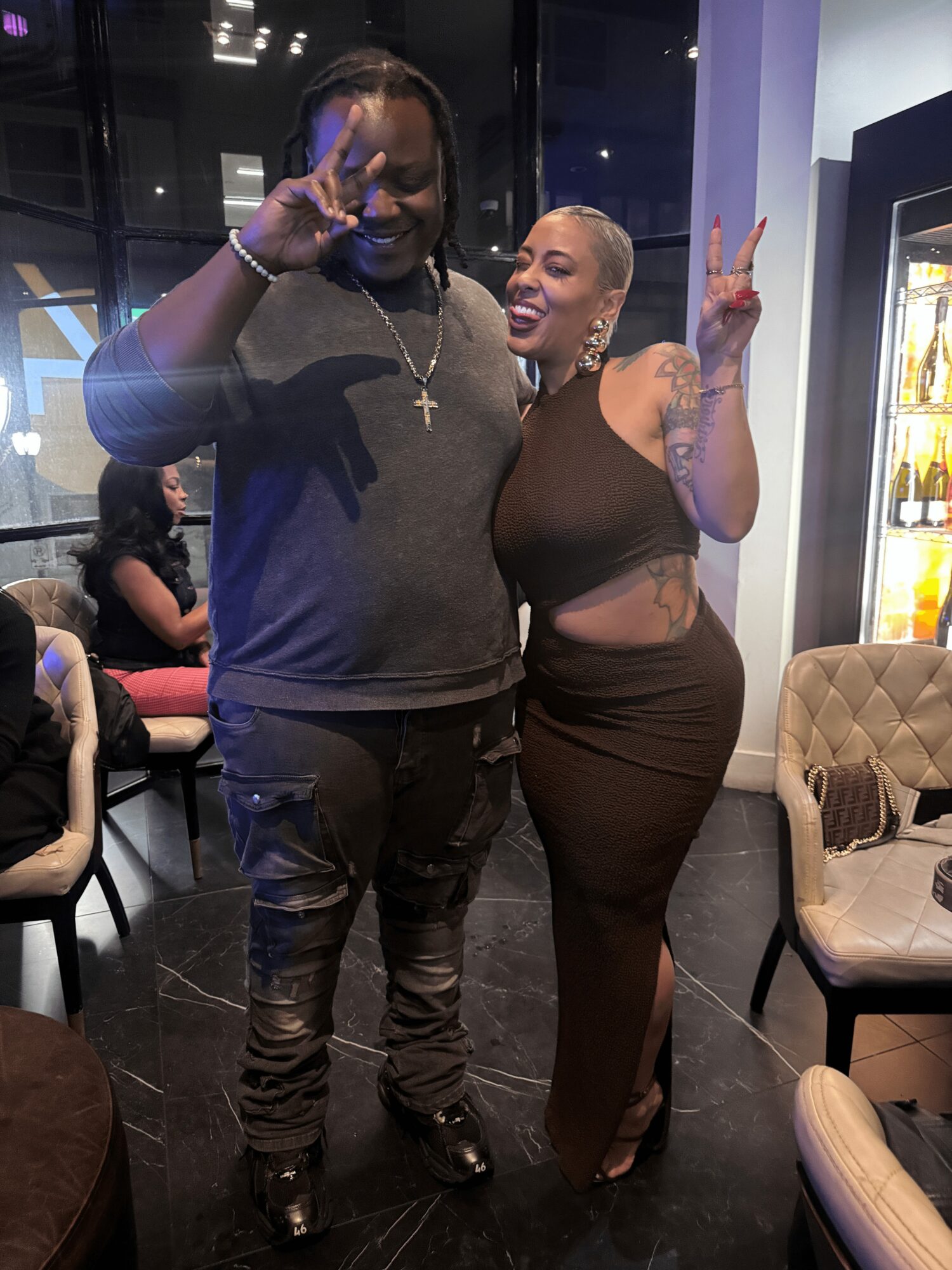 Two people standing indoors, smiling, making peace signs, with chairs and a vending machine in the background.