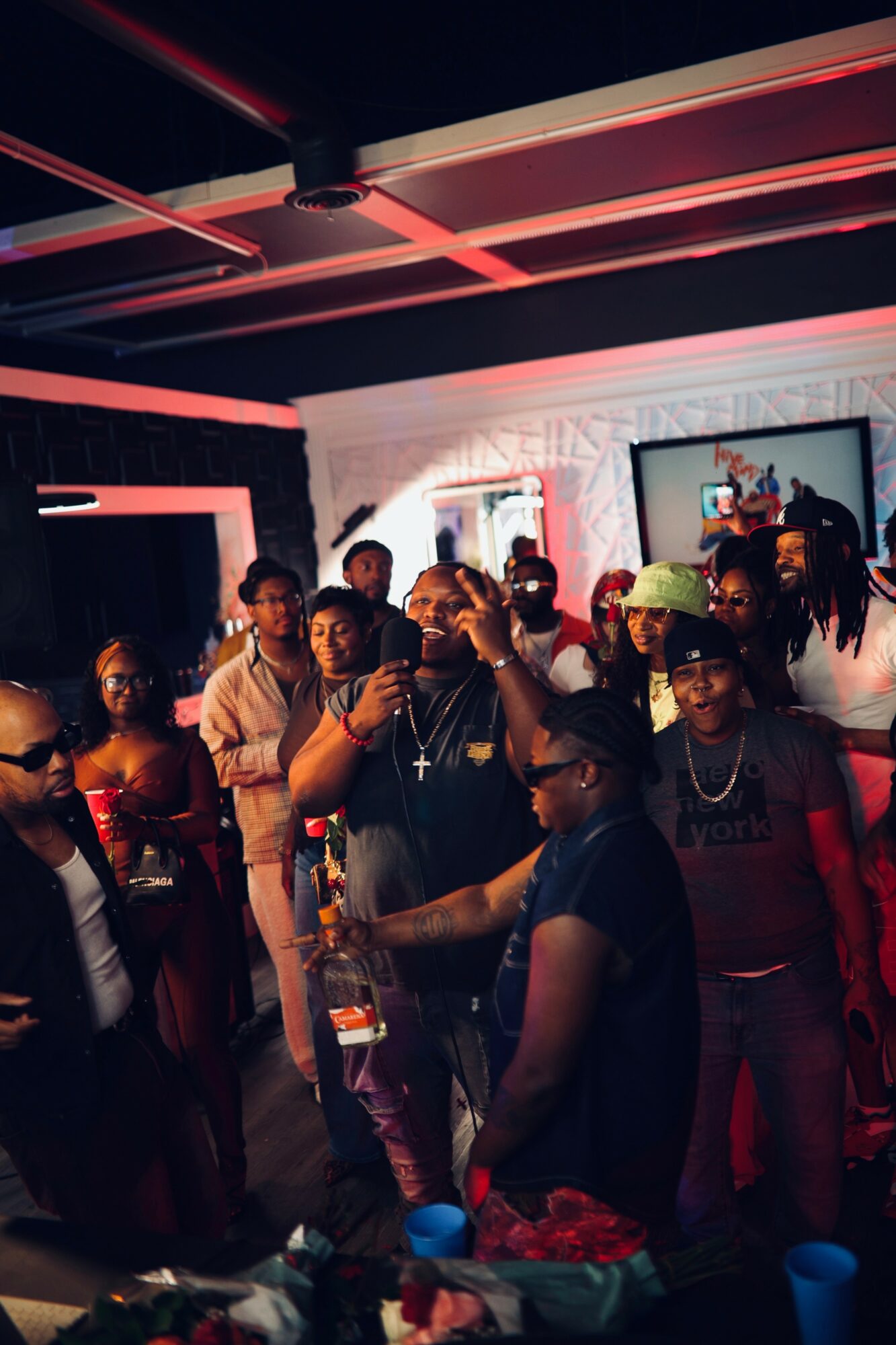 Group of people socializing indoors with neon lighting and wall art, some holding drinks, in a lively setting.