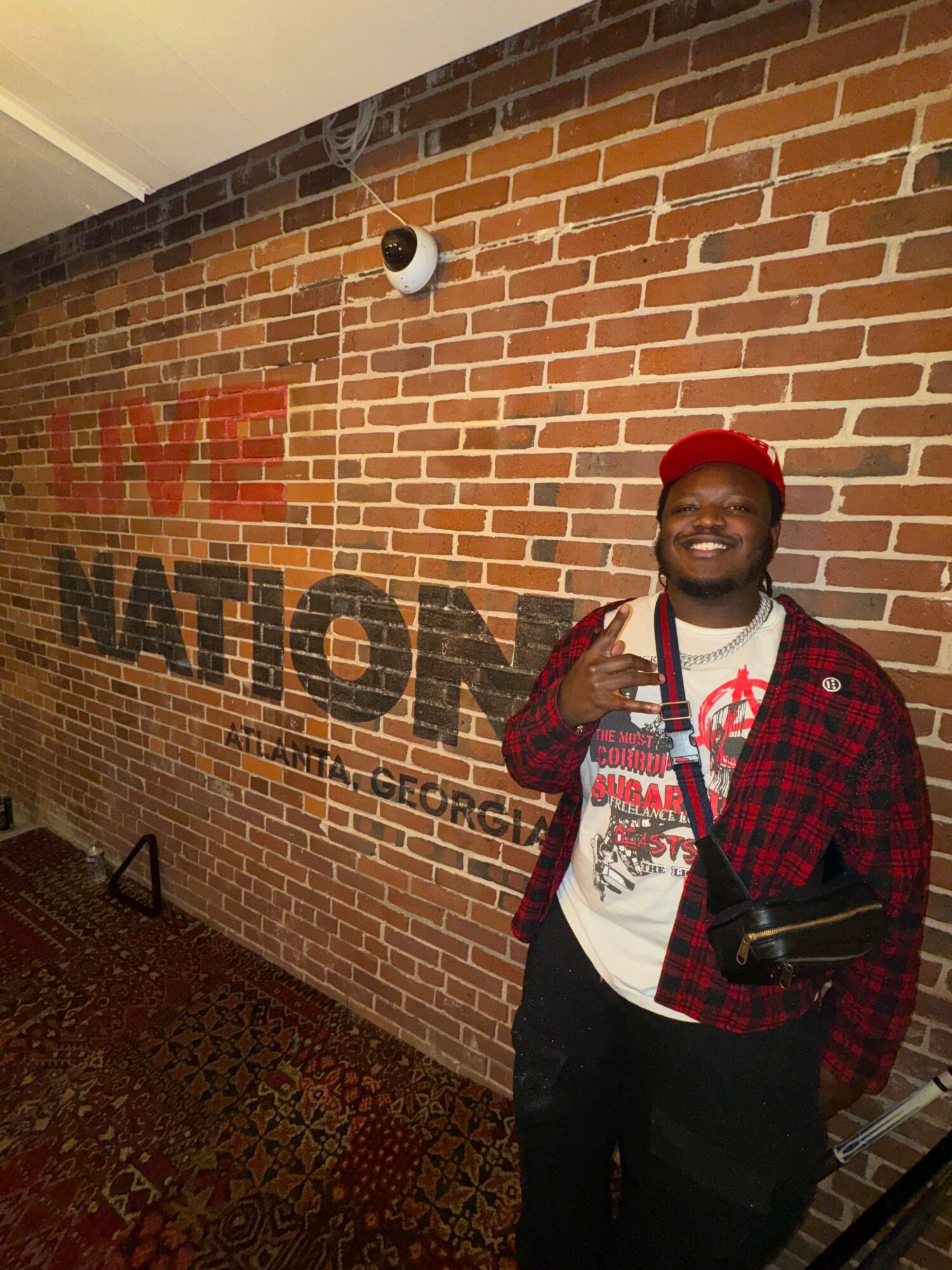 Smiling man wearing a red cap and plaid jacket standing in front of a brick wall with TV and NATION text.