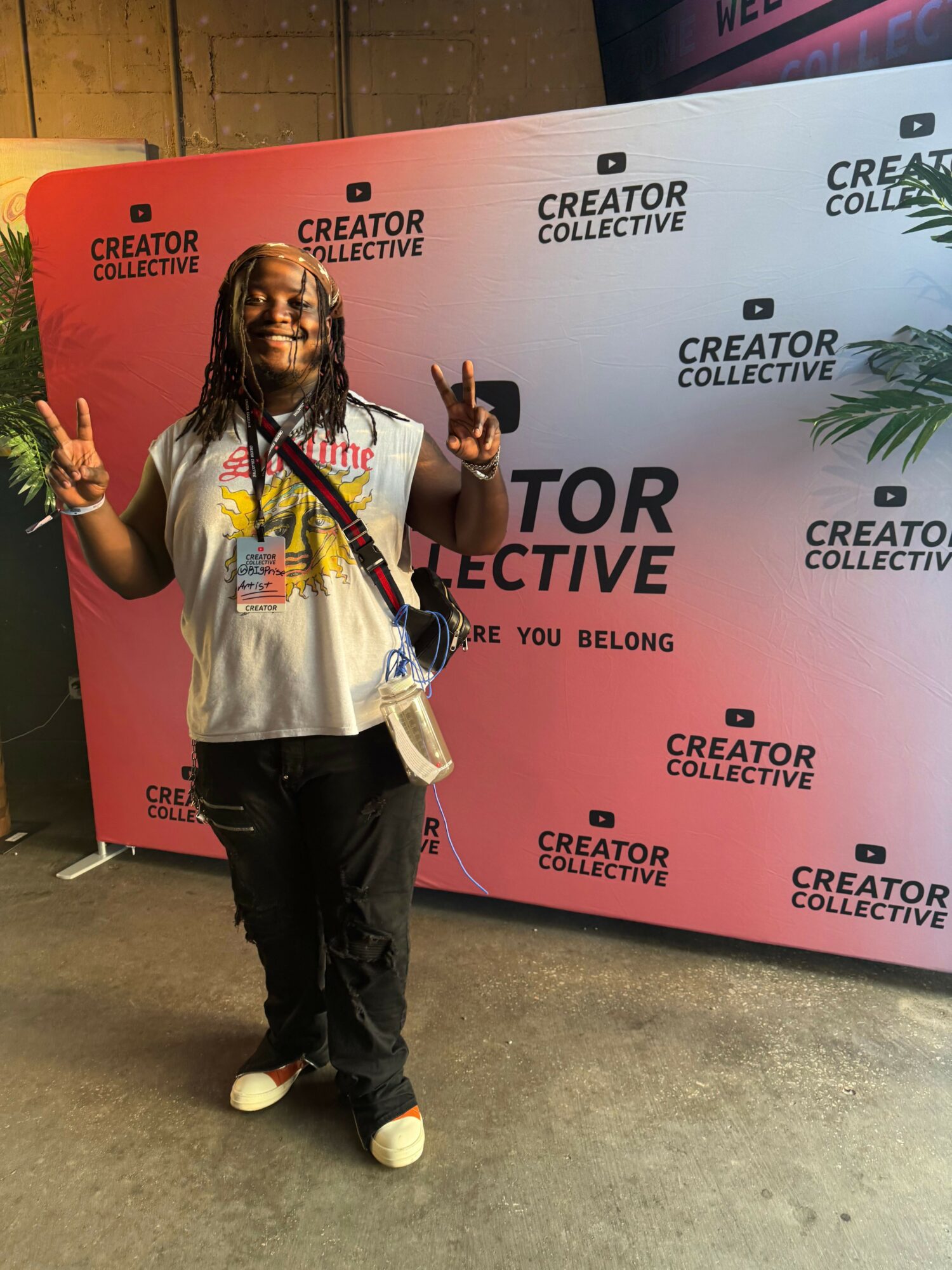Person with long hair and glasses smiling, making peace signs, standing in front of a Creator Collective backdrop.