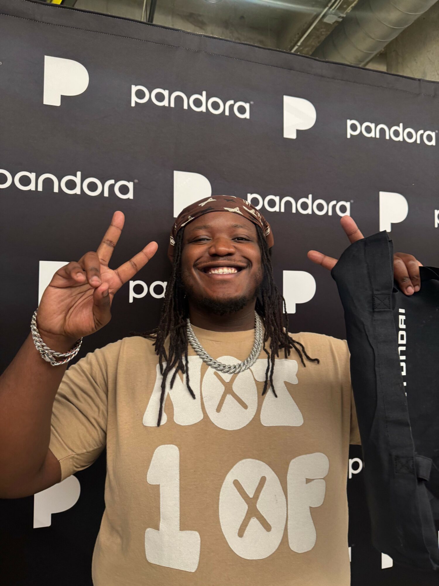 Person smiling, holding peace signs near head, standing in front of Pandora backdrop, wearing beige shirt and jewelry.