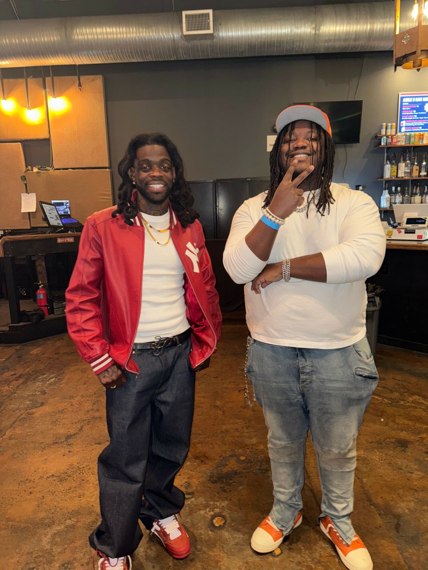 Two men stand indoors, smiling, with bar and TV in background. One wears red jacket, the other white hoodie and cap.