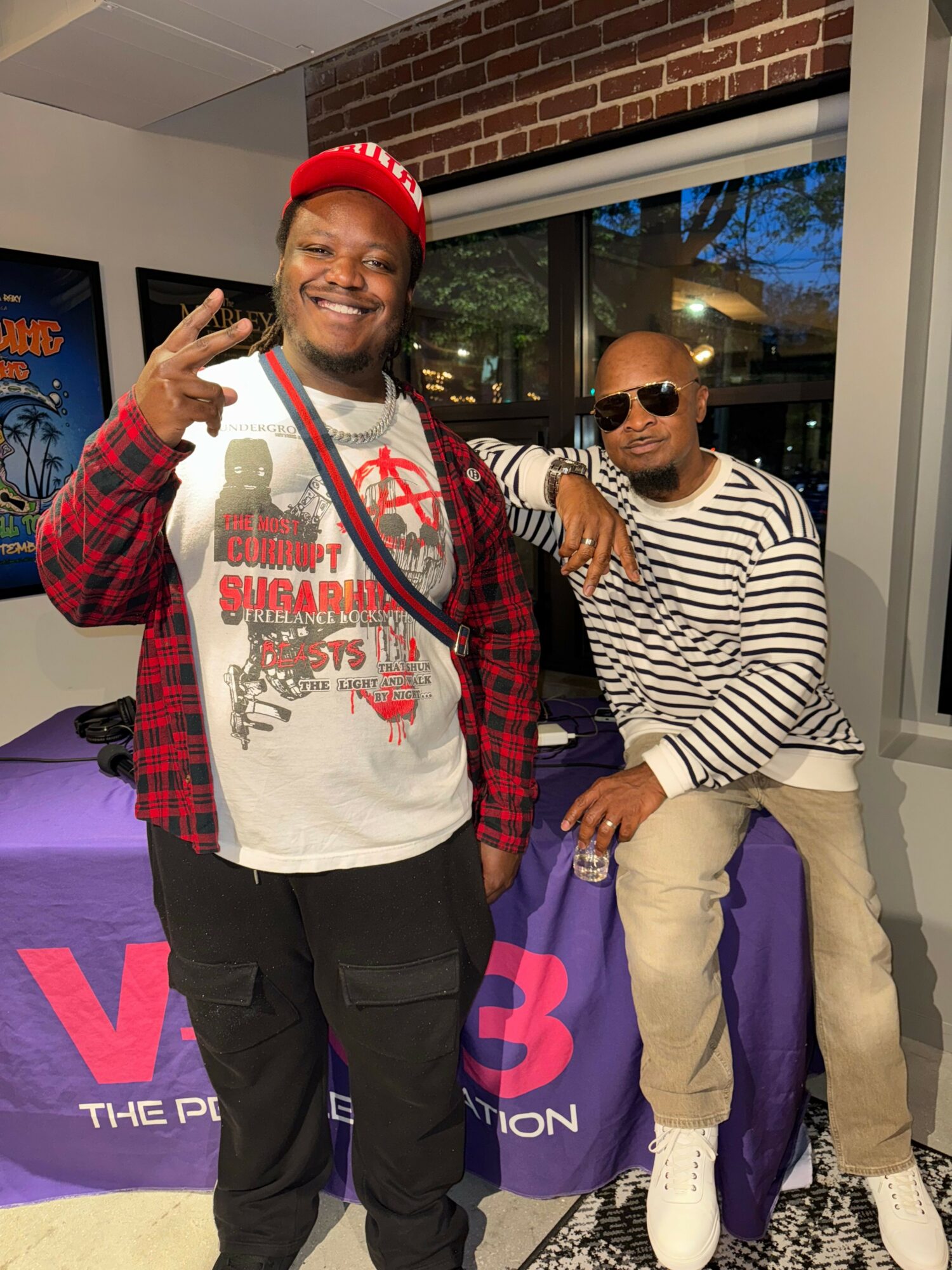 Two people smiling indoors, one wearing a red cap and striped shirt, the other in sunglasses and striped top, near a table.