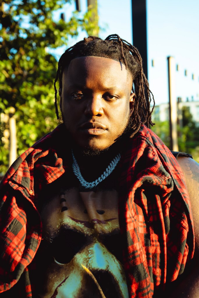 Young man with dreadlocks wearing a red plaid jacket and a chain necklace outdoors.