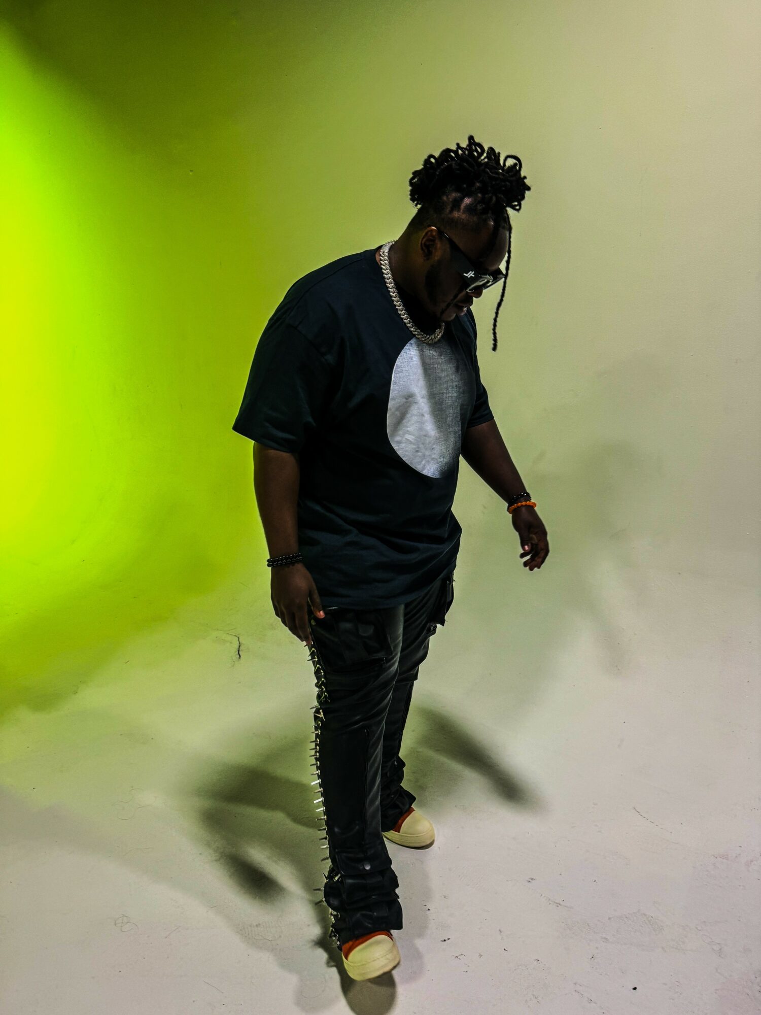 Young man with glasses and dreadlocks wearing a black t-shirt and pants, standing on a white surface.