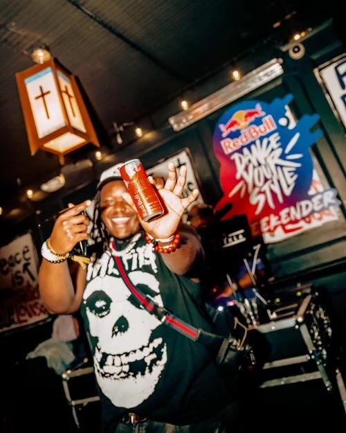 Person smiling, holding a red can, wearing a black T-shirt with a skull design, in a lively indoor setting.