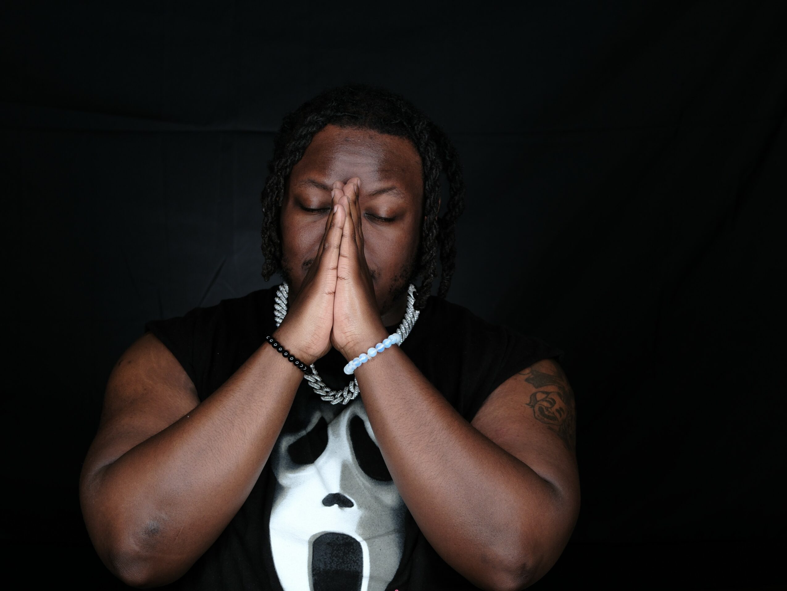 Person with eyes closed, hands pressed together in prayer or reflection, against a dark background.