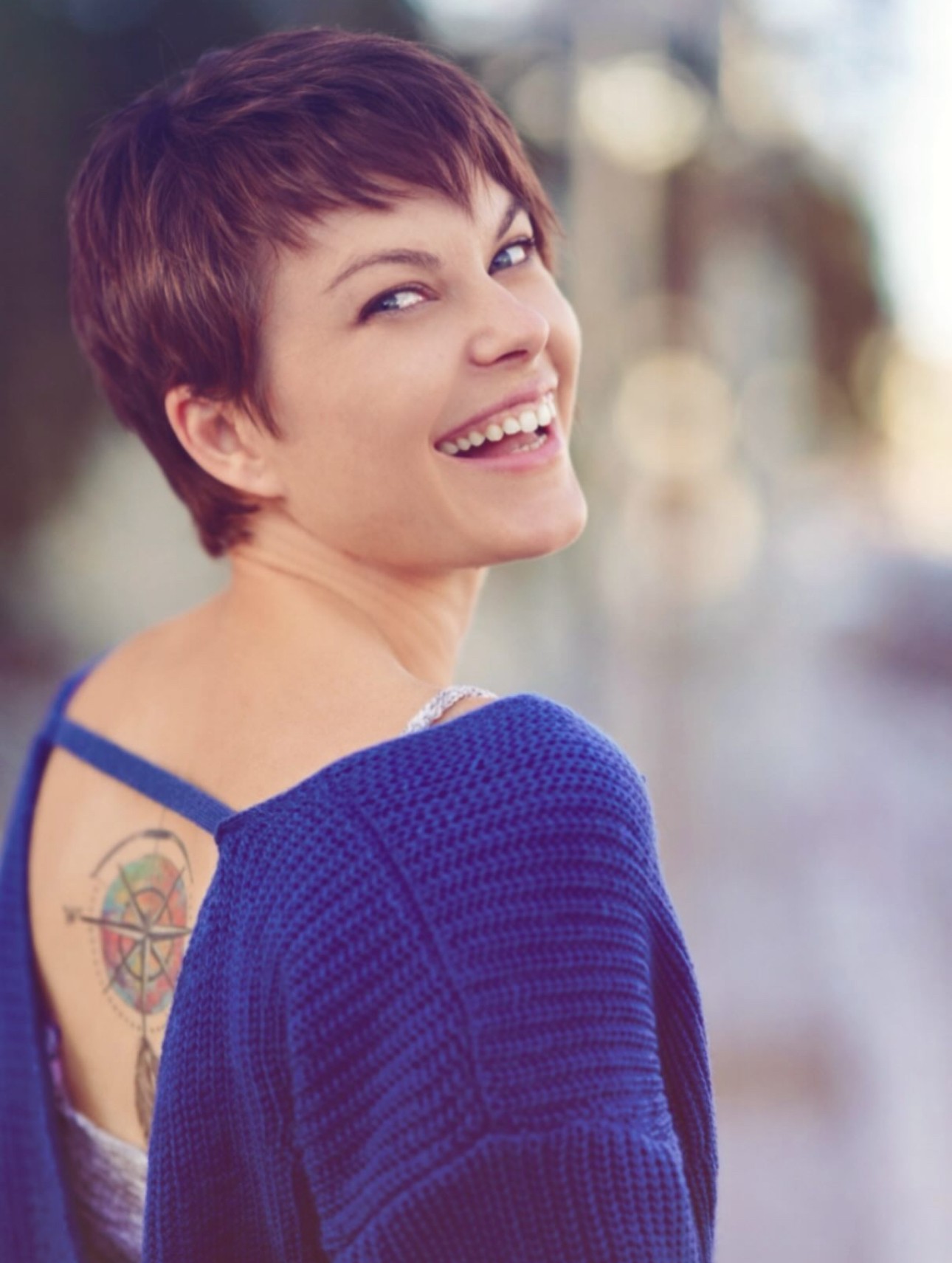 Woman with short hair smiling, wearing a blue sweater, outdoors with blurred background.