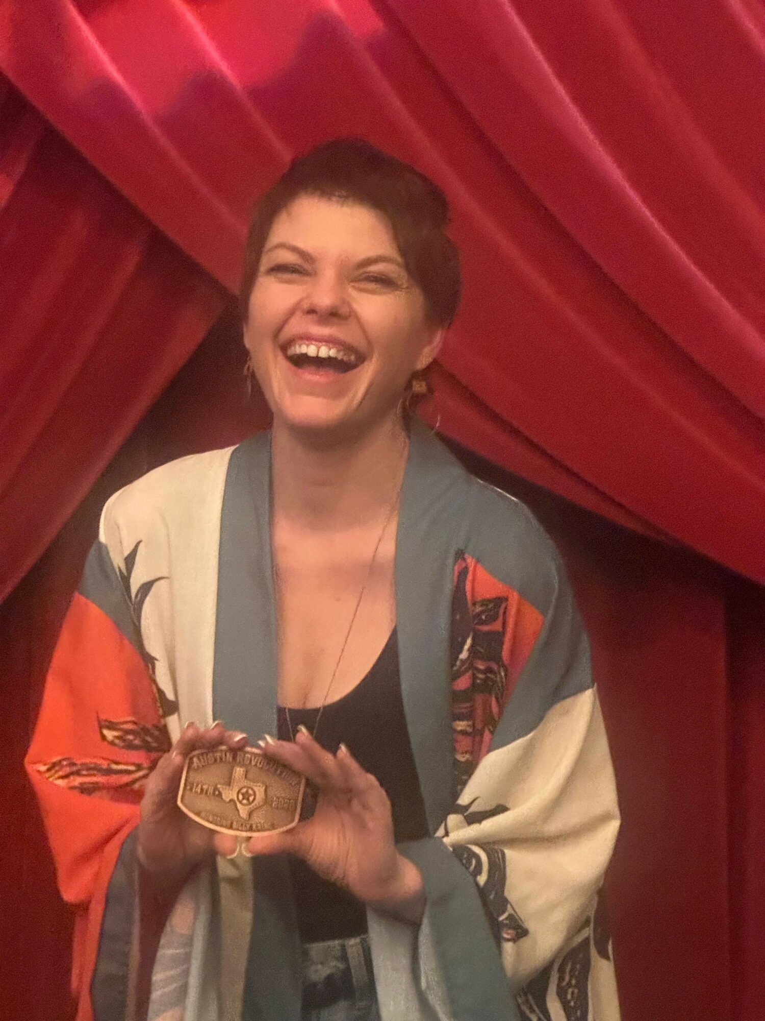 Woman with short hair smiling and holding a small object, standing in front of red curtains.