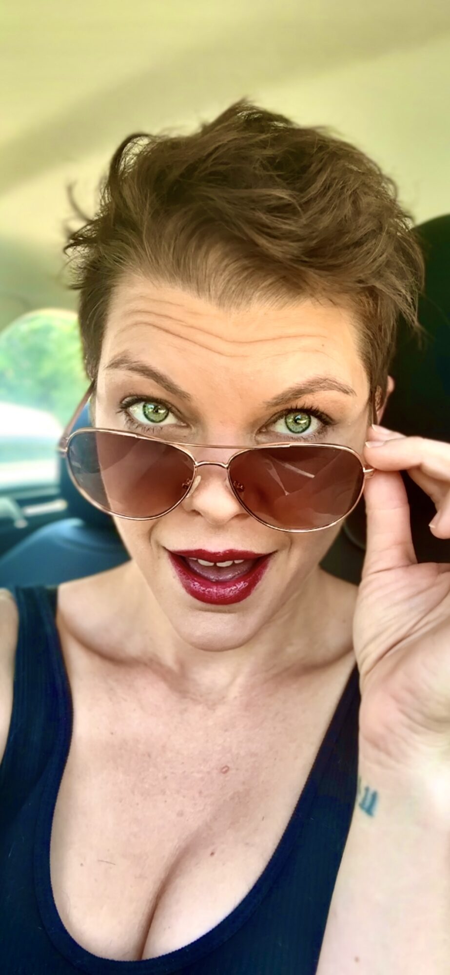 Woman with short hair, wearing sunglasses and a dark top, looking at camera with a surprised expression.