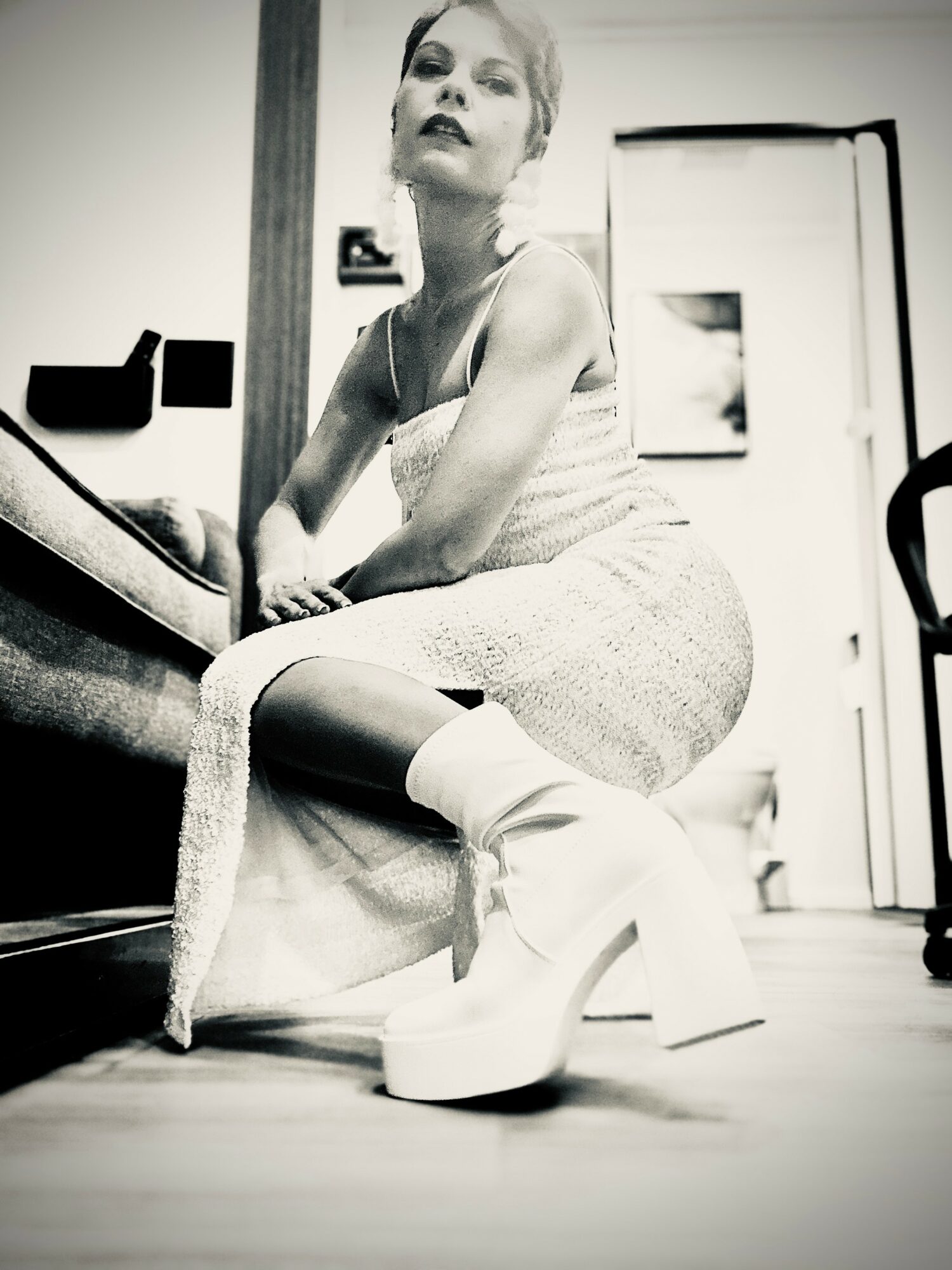 Person sitting on a chair wearing high platform boots, with a dress and short hair, in a room with a door and mirror.