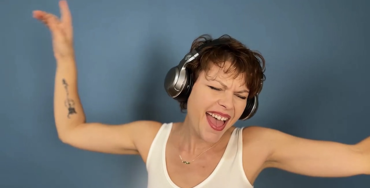 Woman with short hair wearing headphones, smiling with arms raised, against a blue background.