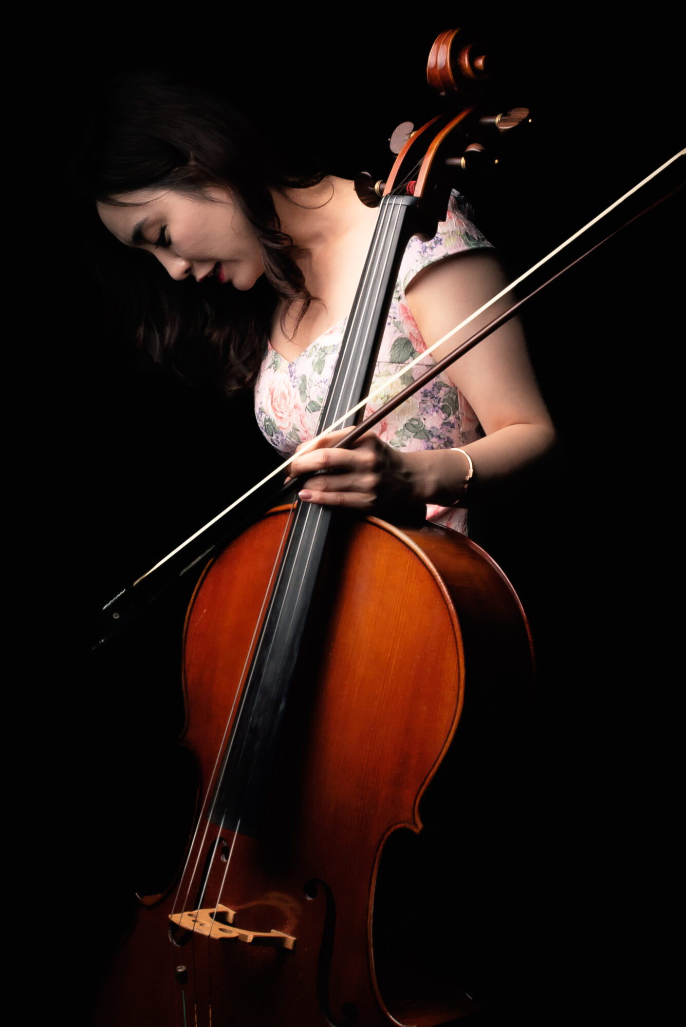 Woman playing a cello with a bow, dark background, focused expression, wearing a floral dress.