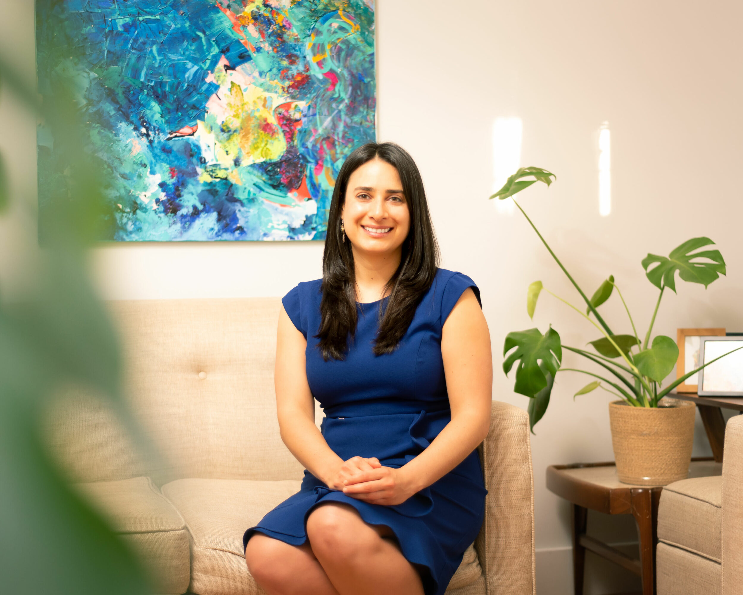 Woman sitting on a beige sofa, smiling, wearing a blue dress, with a colorful abstract painting behind her.