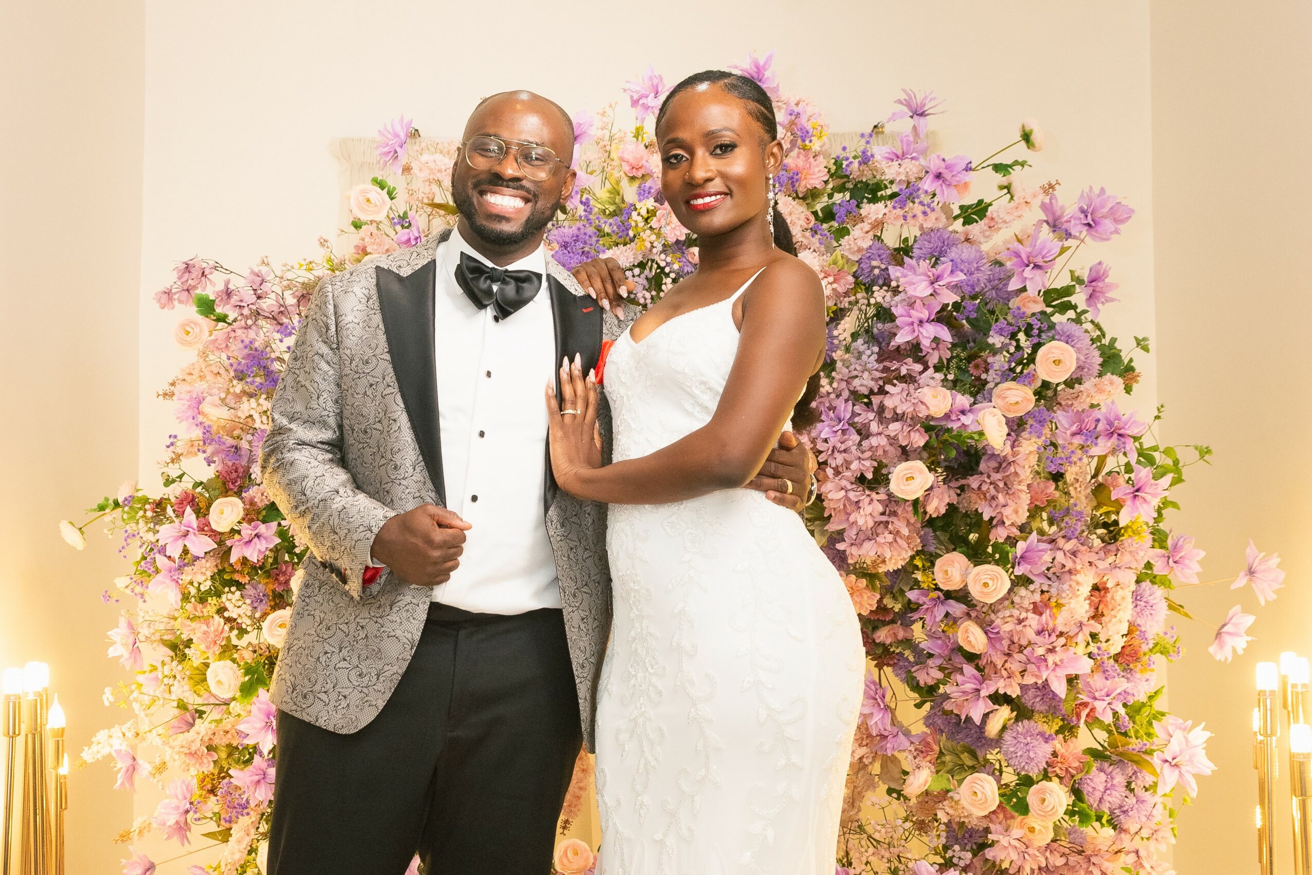 A smiling man and woman in formal attire stand in front of a large floral arrangement with pink and purple flowers.