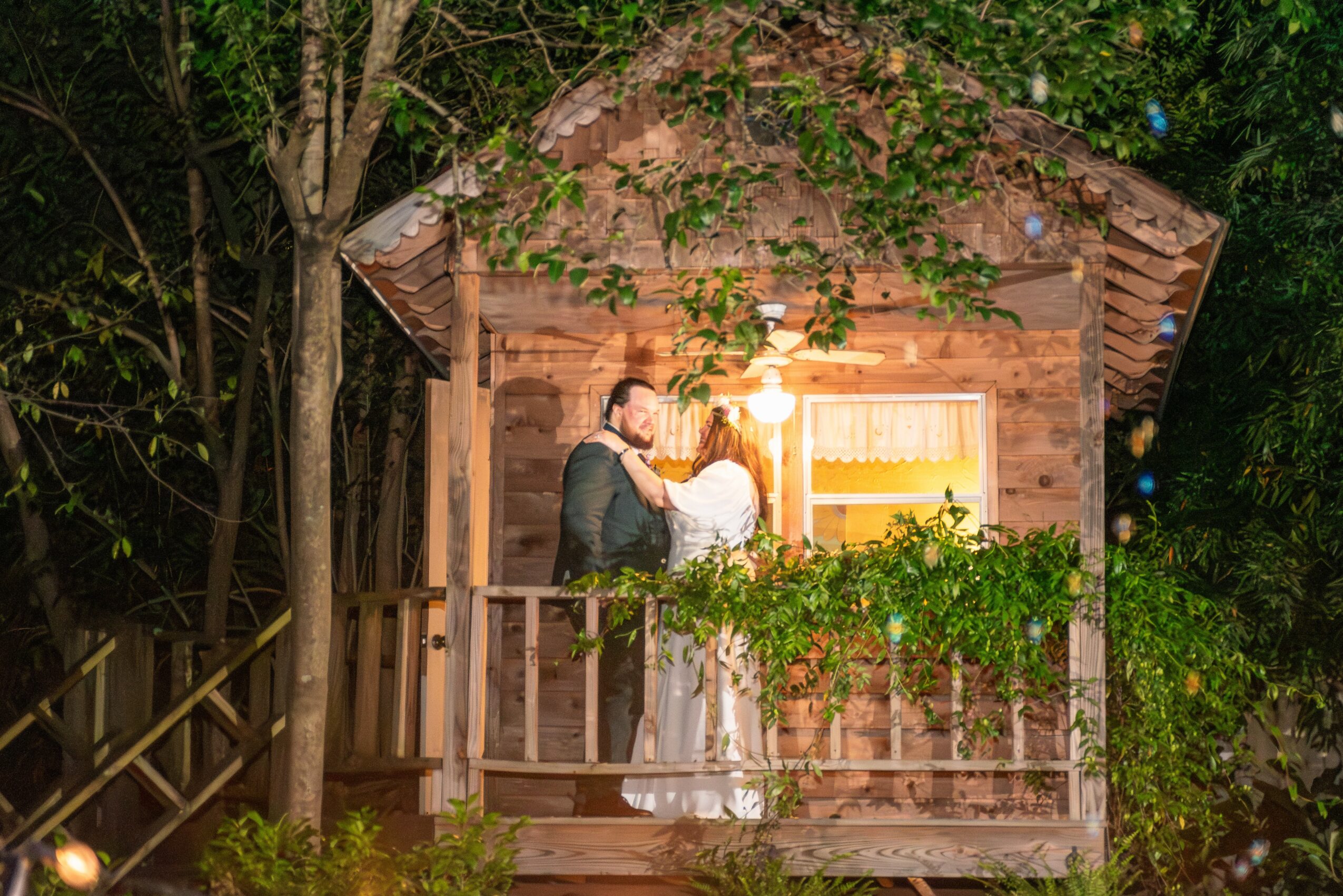A couple stands on a wooden porch in front of a small house surrounded by trees and greenery.