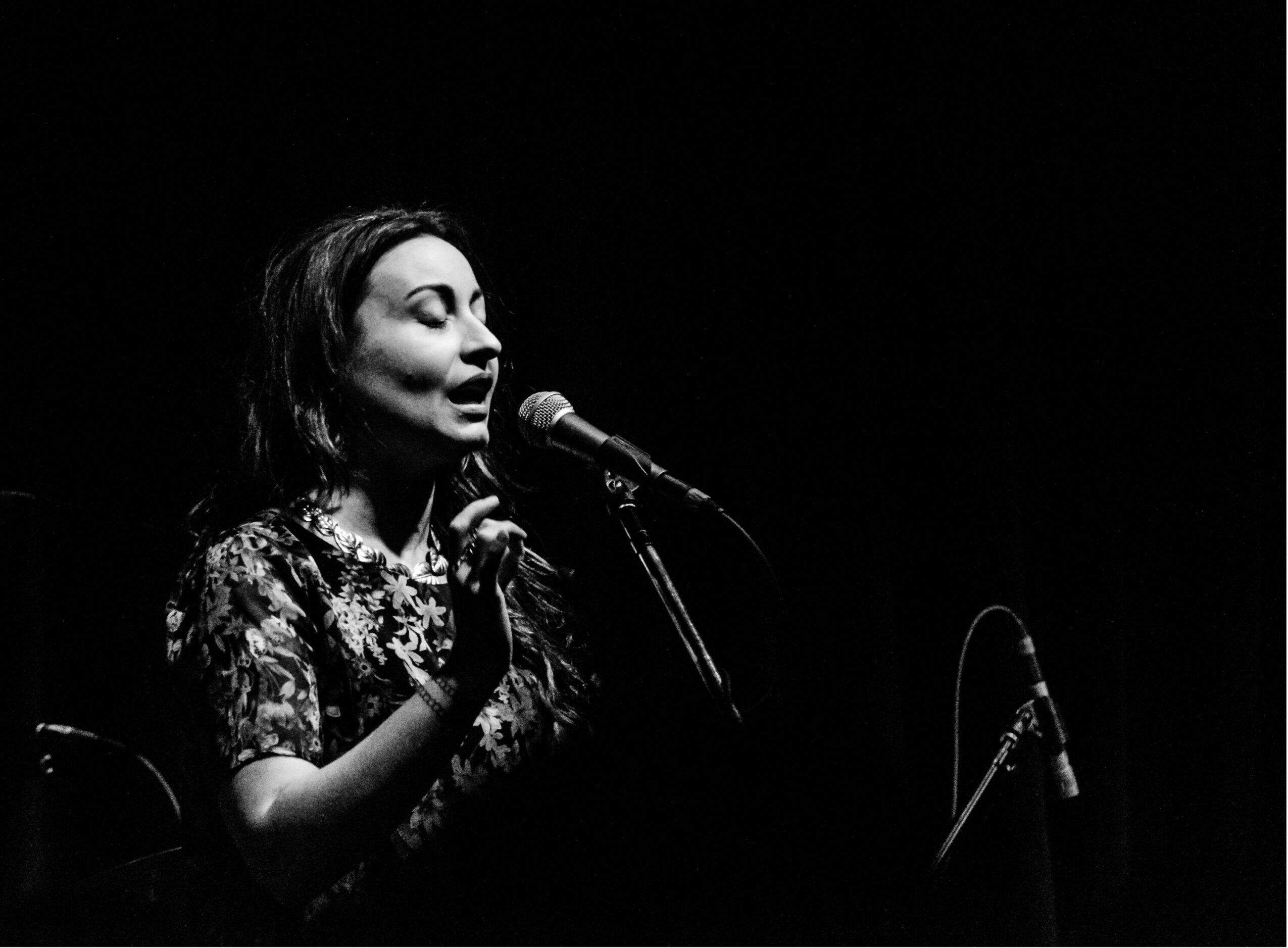 Woman singing into a microphone on stage with a dark background.