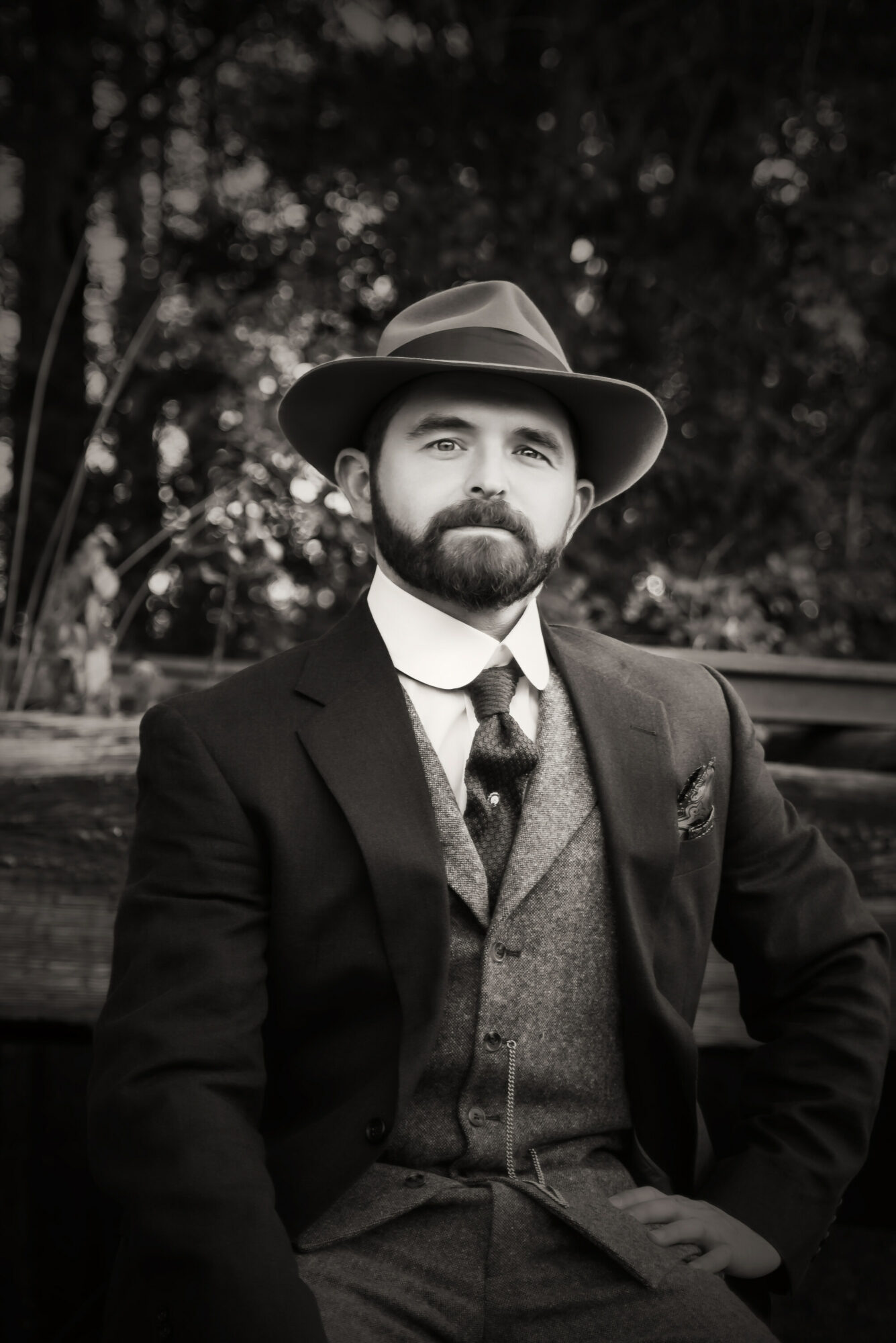 Man with beard in vintage suit and hat outdoors, trees in background.