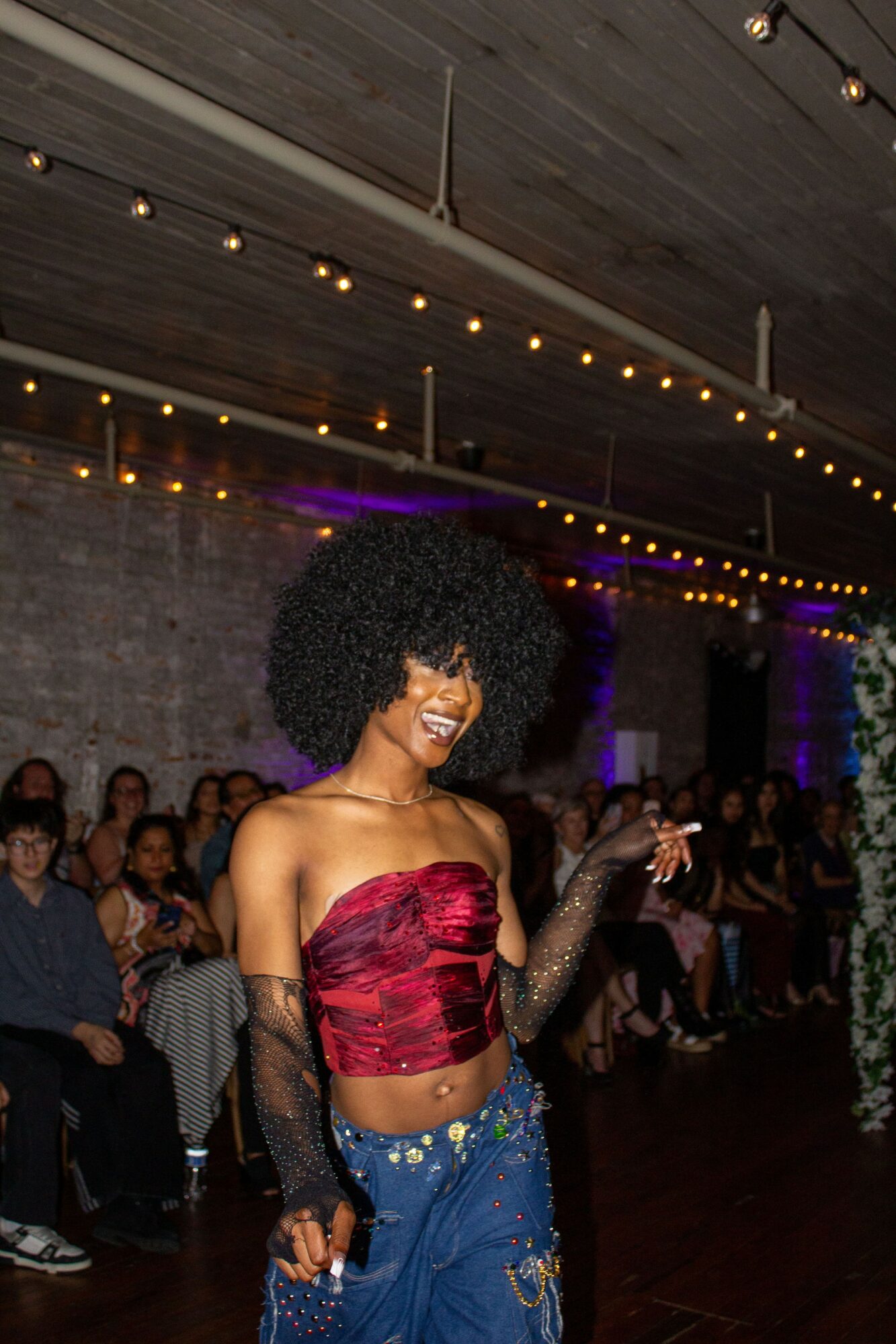 Woman with curly hair in a red top and blue jeans, smiling at a party with string lights and seated audience.