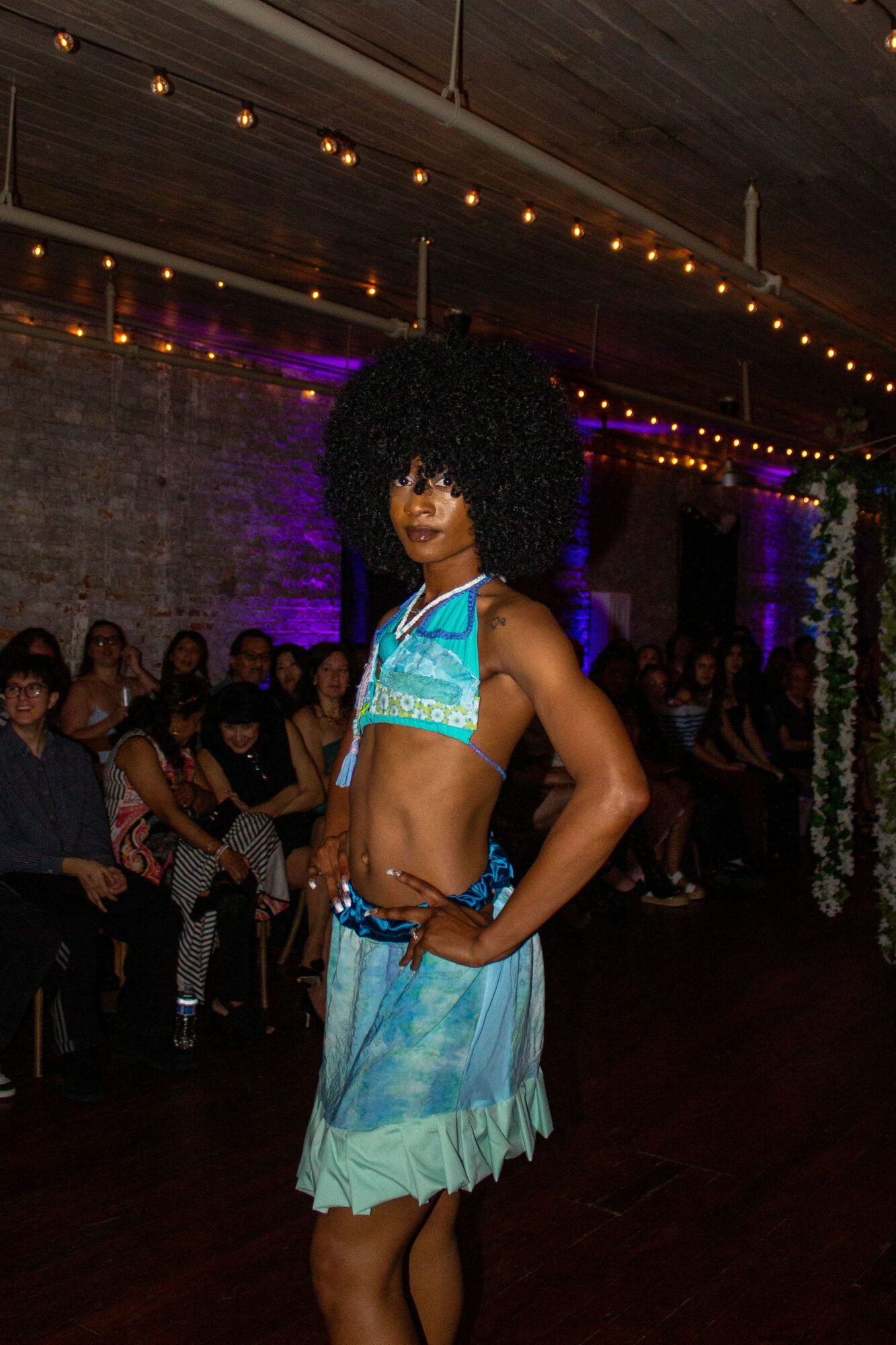 Person with curly hair in a turquoise and white outfit standing in a dimly lit venue with seated audience.