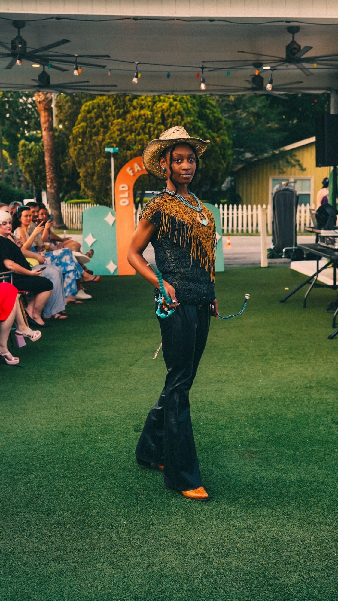 Woman standing on grass in stylish outfit with hat, surrounded by seated people and outdoor setting.