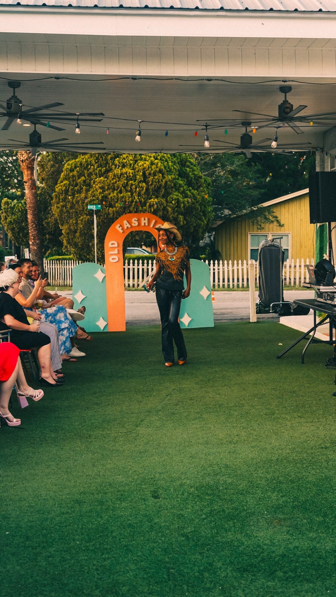 Person standing in front of a colorful arch at an outdoor event, with seated audience on the left and trees in background.