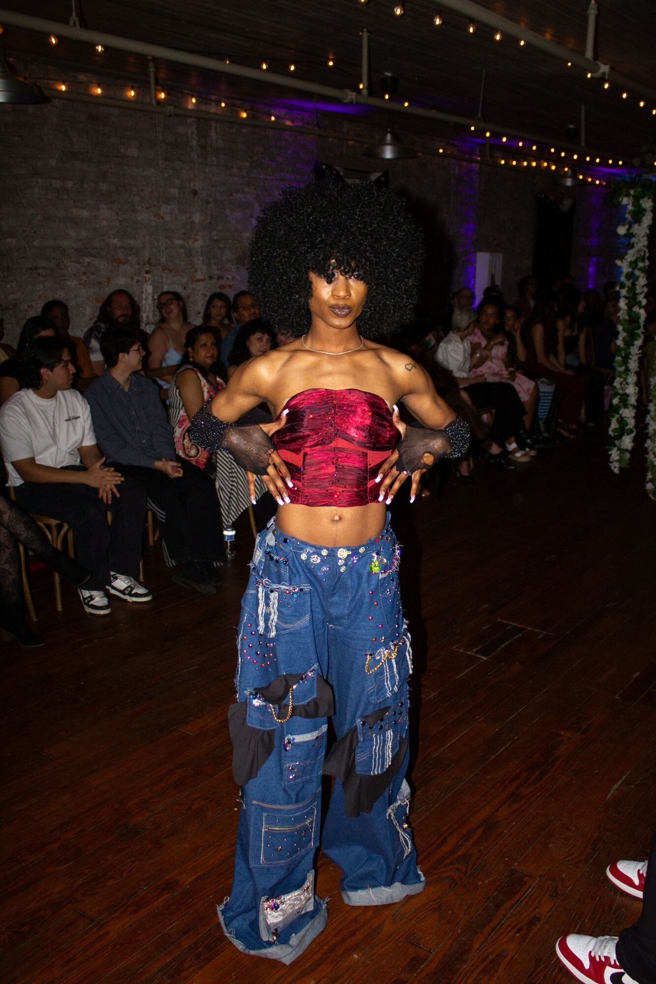 Person with large curly hair wearing a red top and baggy blue jeans in a room with seated audience.