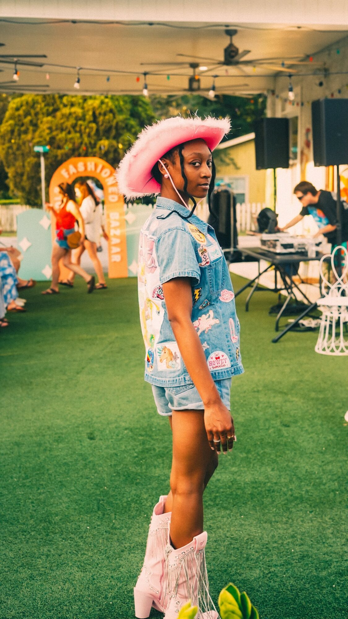 Young girl in denim outfit and pink hat standing outdoors at event with people and decorations.