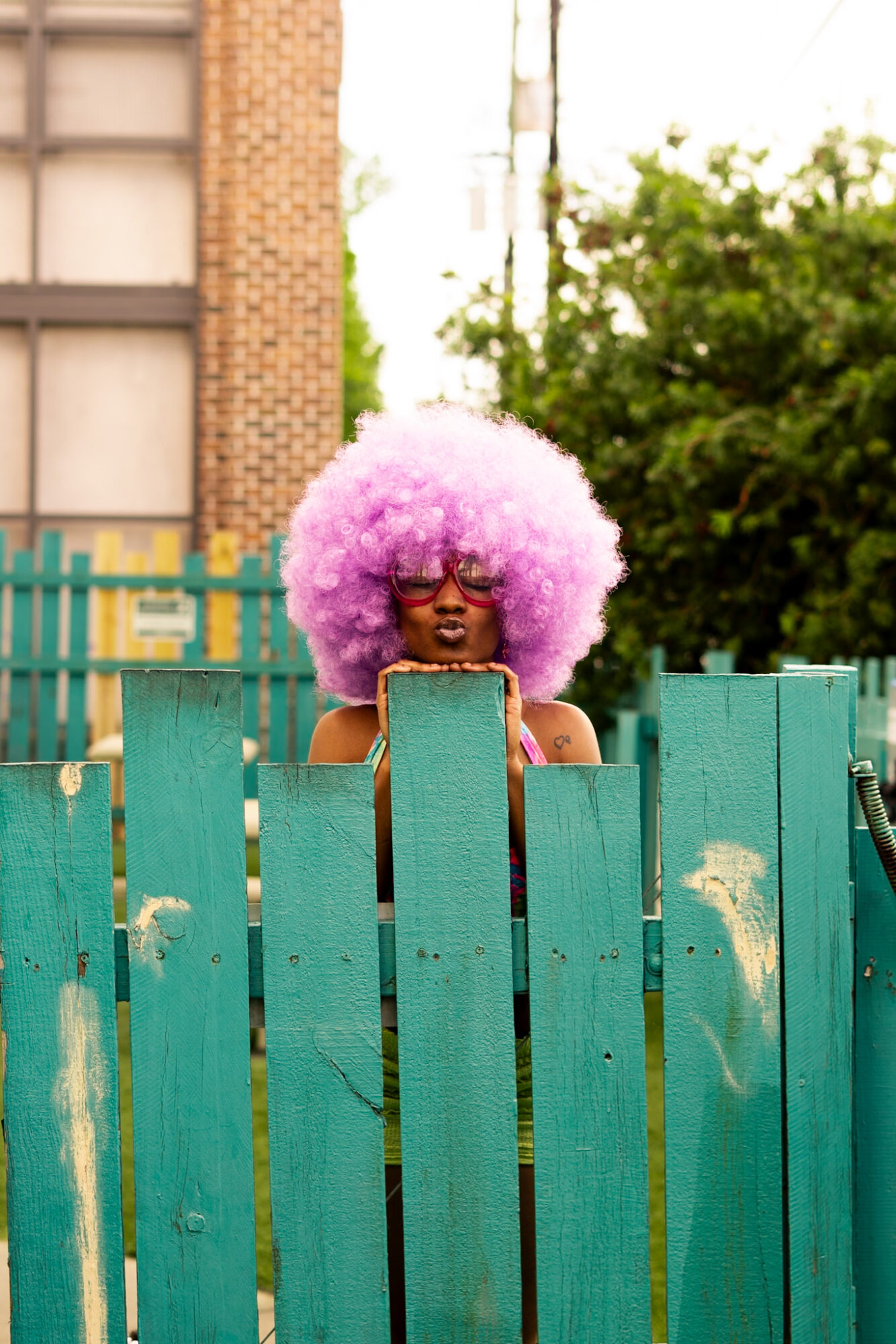 Person with large pink curly wig and sunglasses peeks over a teal wooden fence outdoors.