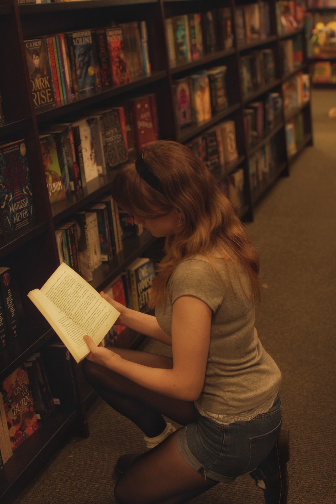 Young woman with long hair reading a book in a bookstore aisle, shelves filled with books behind her.