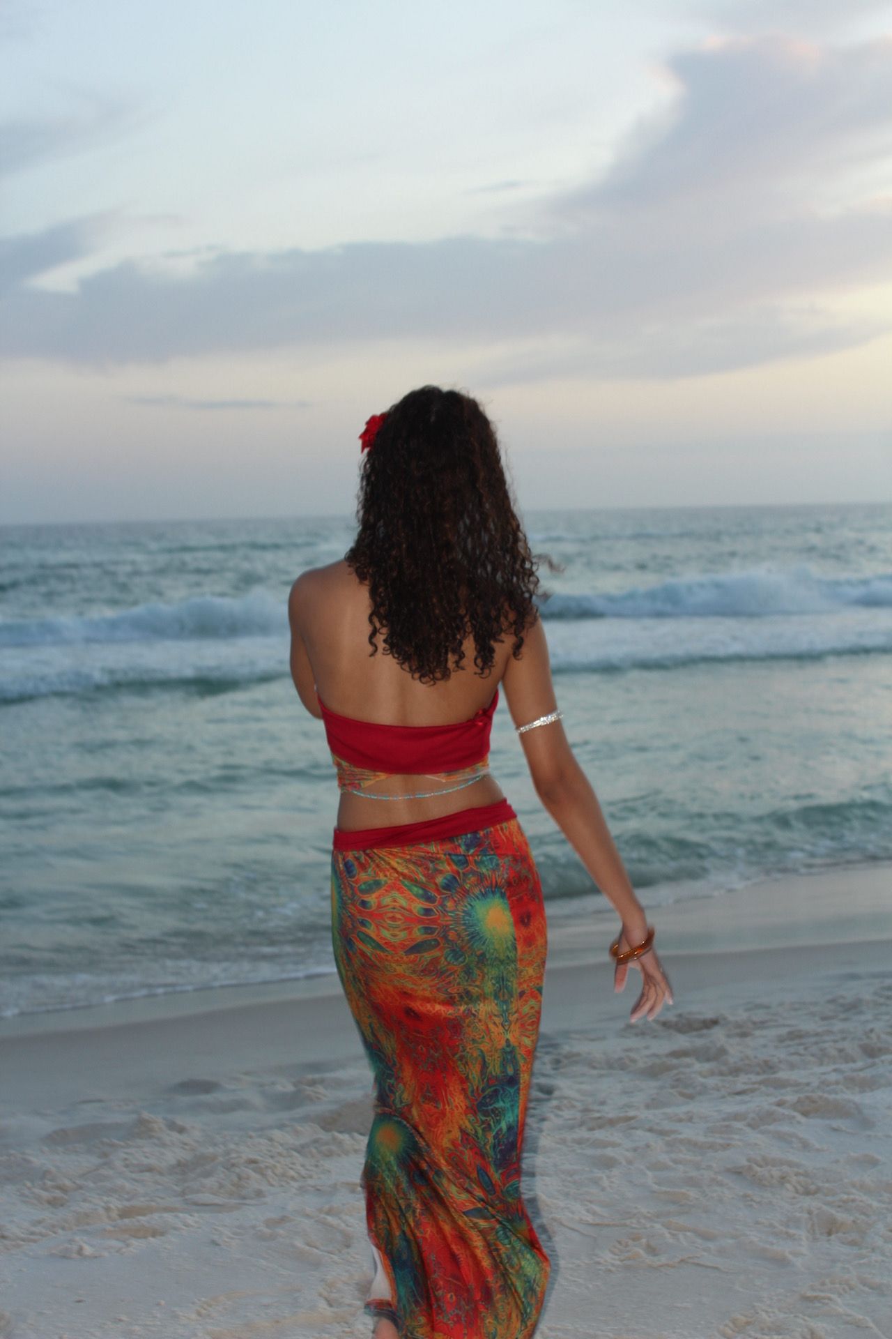Woman with curly hair in a red top and colorful skirt standing on the beach facing the ocean.