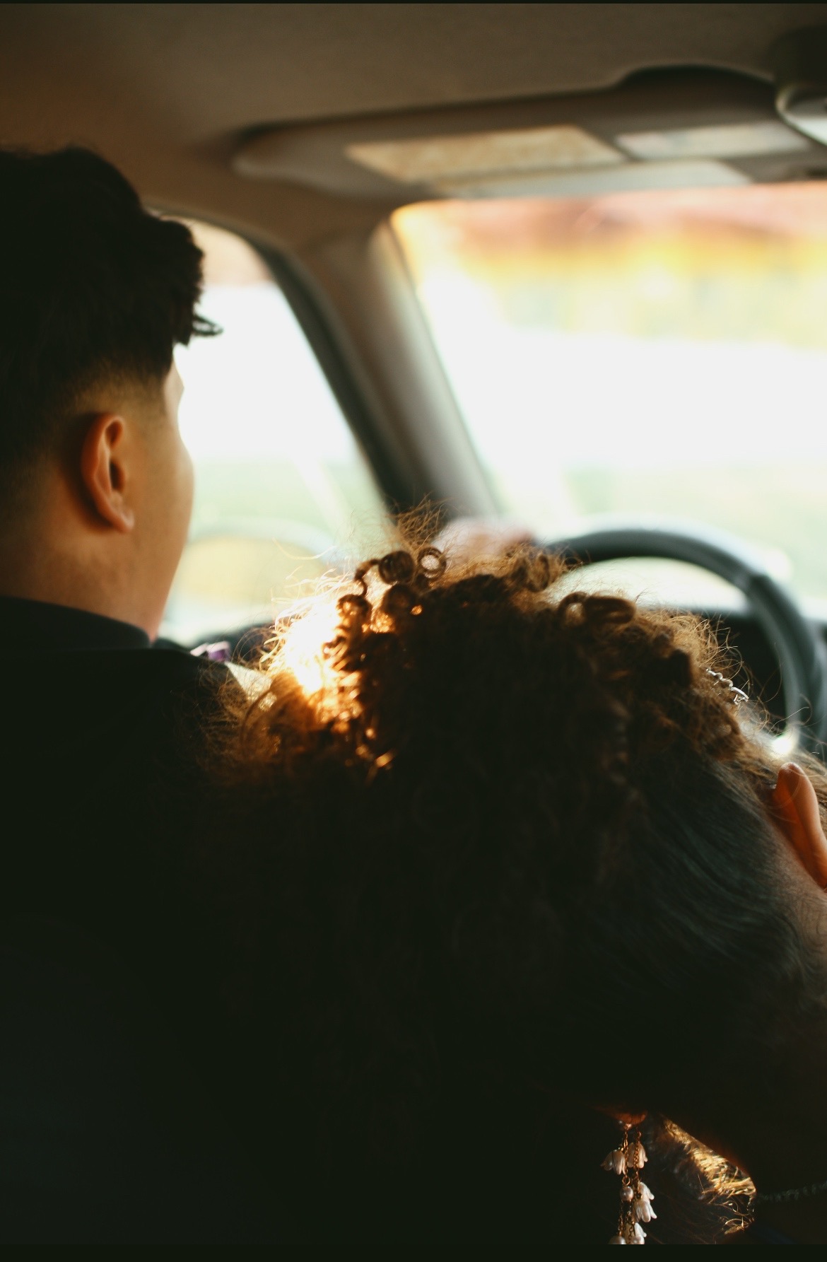 Two people sitting in a car, one driving and the other resting their head on the driver's shoulder, with sunlight outside.