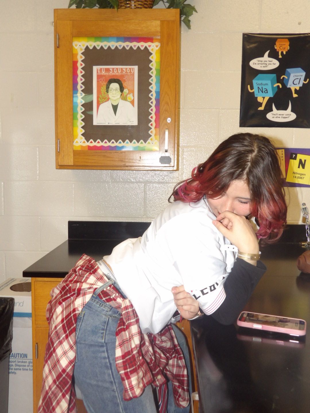 Person with shoulder-length hair, wearing a white shirt and plaid jacket, leaning on a black counter, smiling with eyes closed.