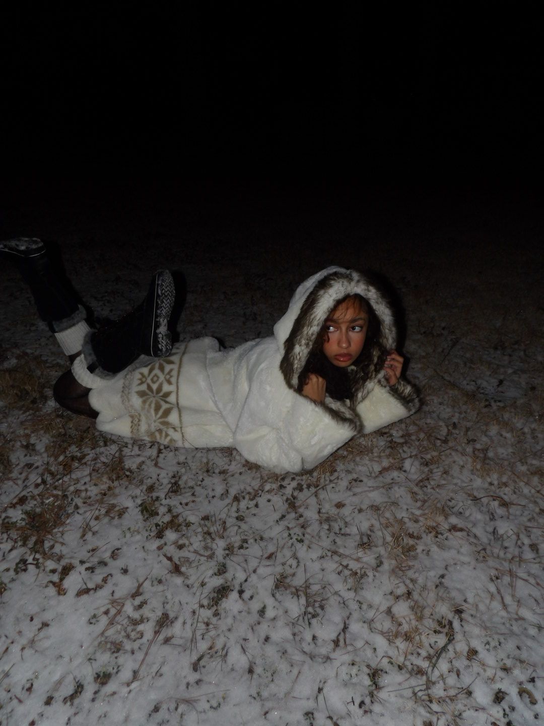 Child lying on snow-covered ground at night, wearing a white hooded coat and patterned pants.