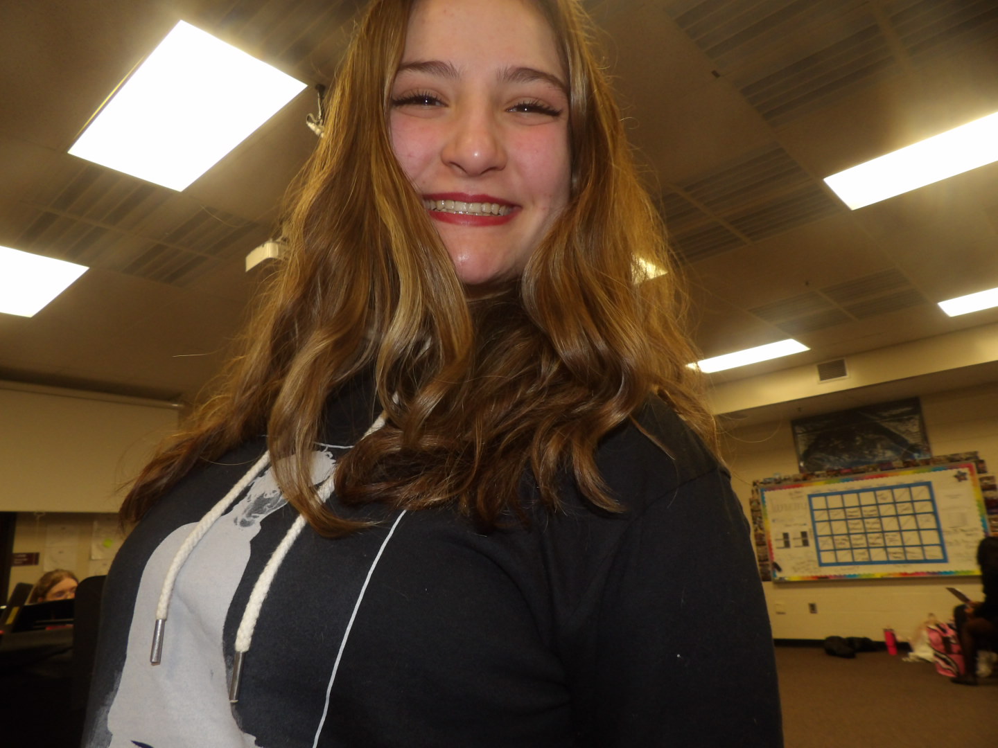 Smiling young woman with long wavy hair in an indoor room with ceiling lights and a whiteboard.