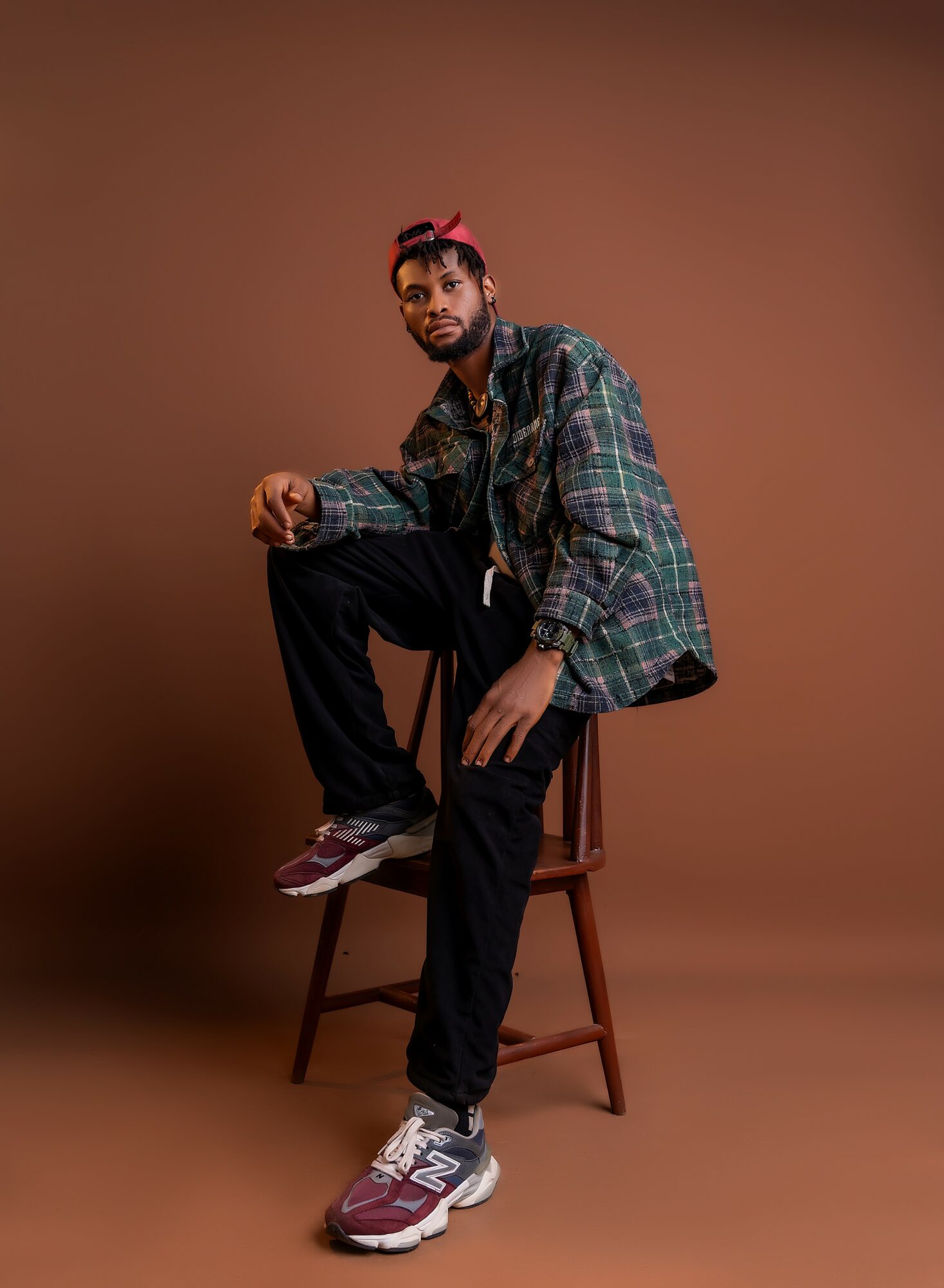 Person sitting on a chair with one leg raised, wearing a plaid jacket, black pants, and sneakers, against a brown background.