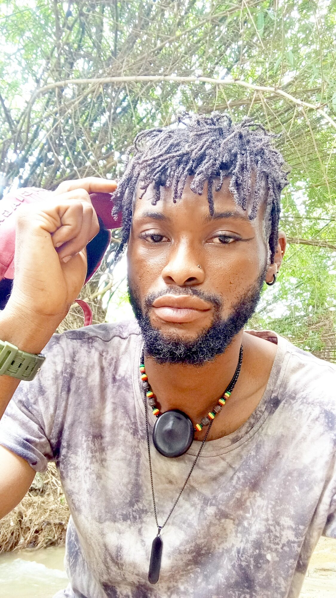 Young man with dreadlocks and beard outdoors, wearing a tie-dye shirt and necklace, holding his head with one hand.
